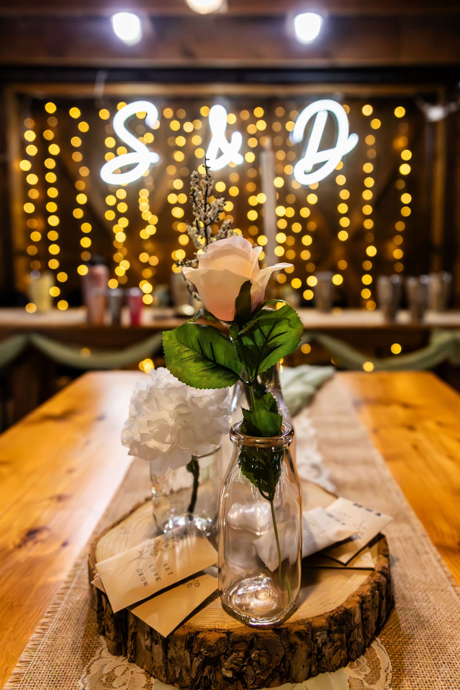 Rustic wedding centerpiece with white rose in glass vase on wood slice with glowing string lights in background.