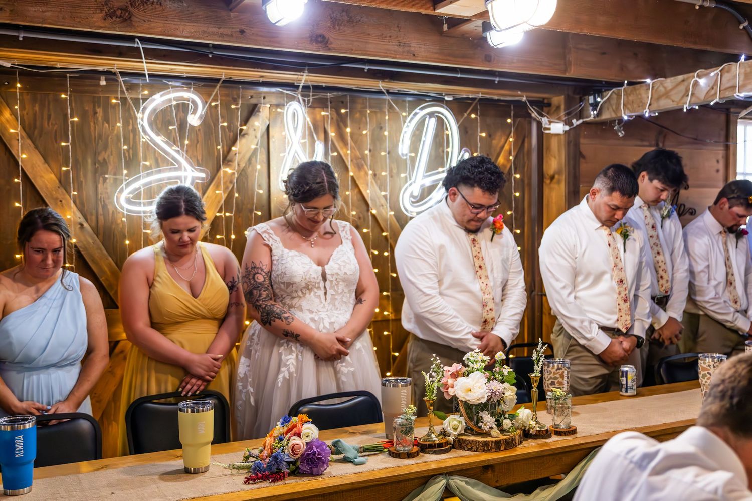 A wedding party gathers at a rustic wooden table decorated with lights and floral centerpieces during a reception.