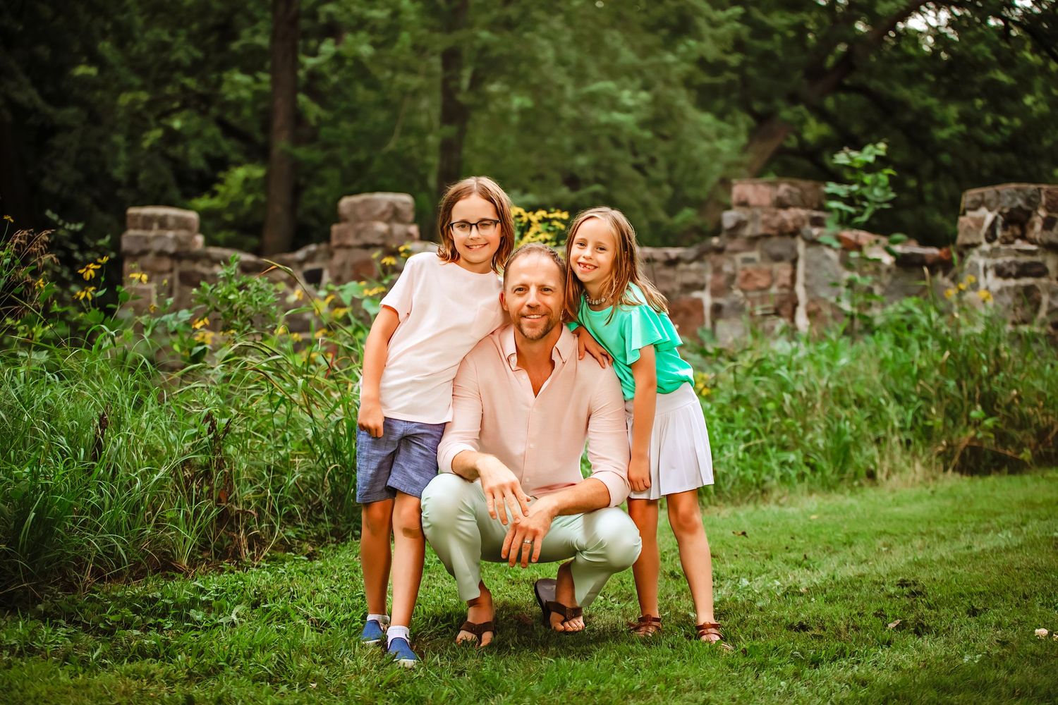 A happy family poses together outdoors on a grassy lawn near an old stone wall on a summer day. Bridge in background in Montevideo, MN.