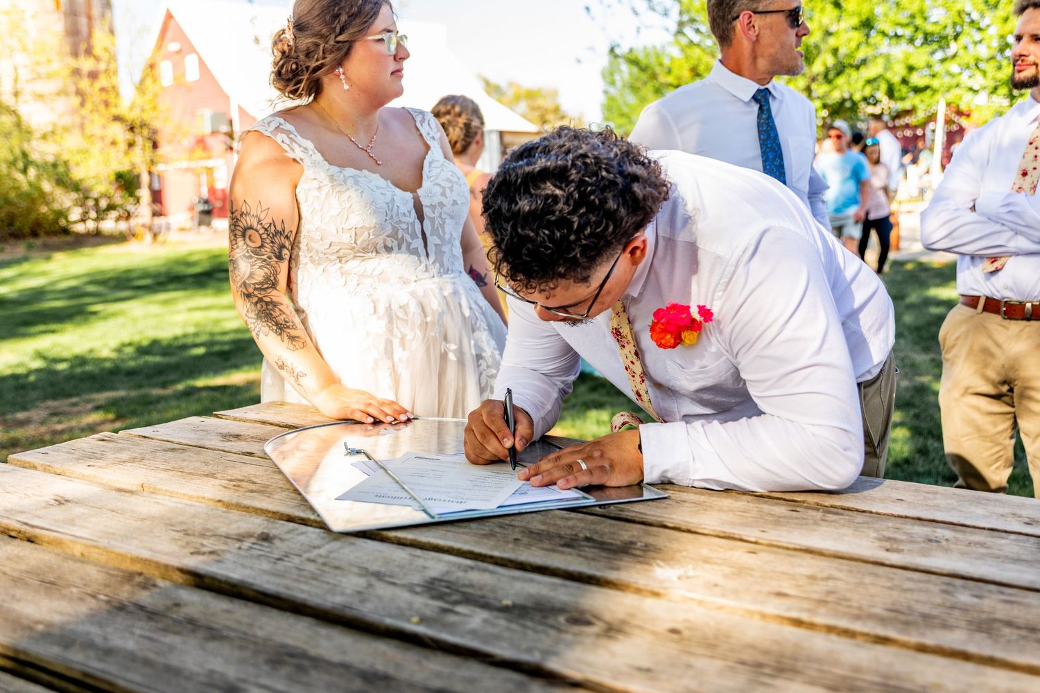 Wedding celebration with guests seated at rustic picnic tables in a natural outdoor setting.