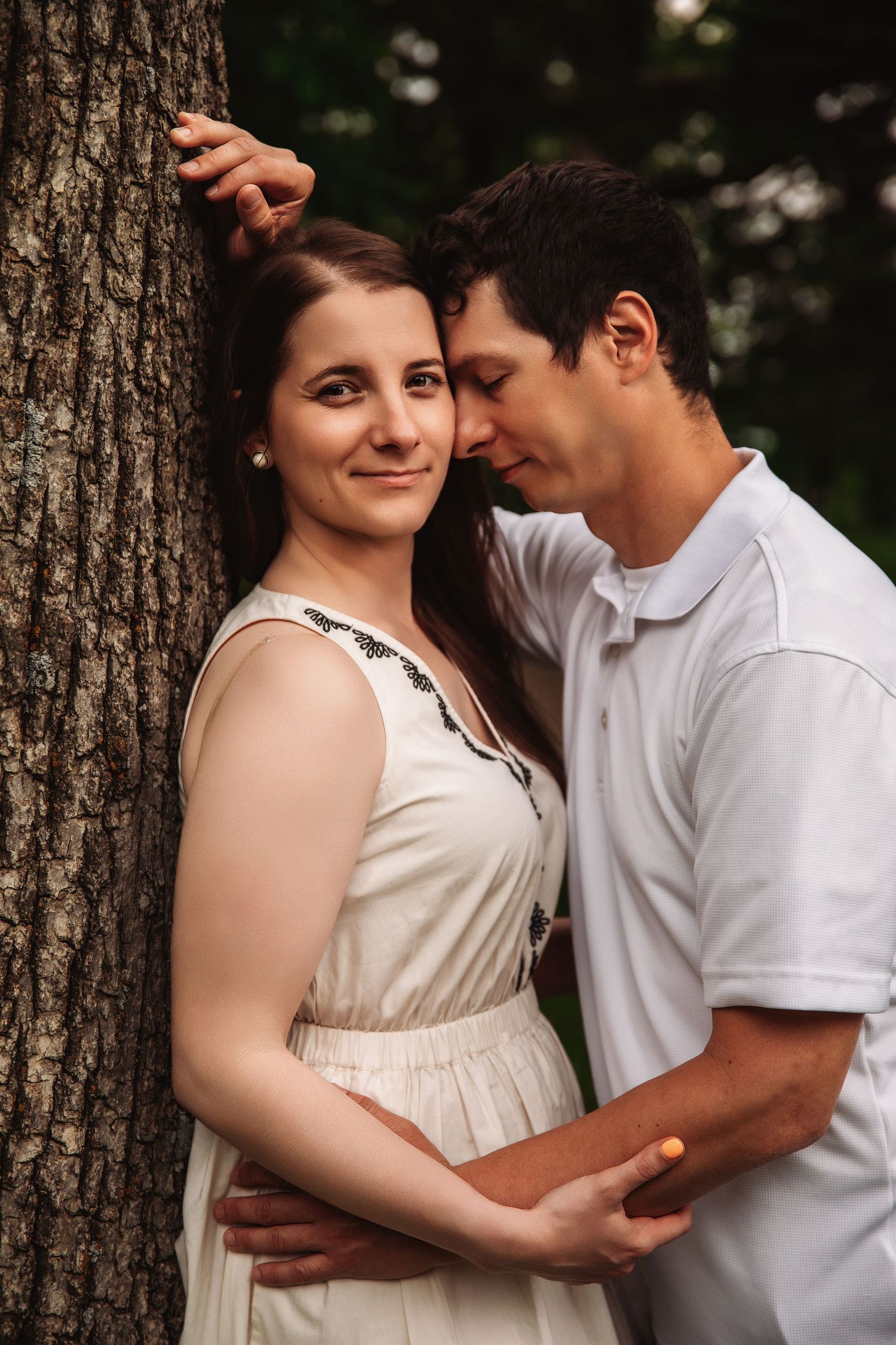 A romantic maternity photo session shows a couple embracing against a tree trunk in natural outdoor lighting.