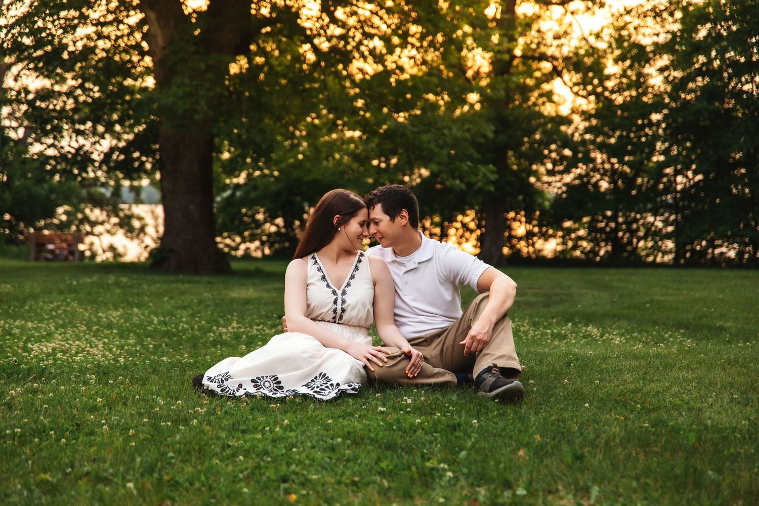 A romantic couple relaxes on lush grass during sunset beneath trees.