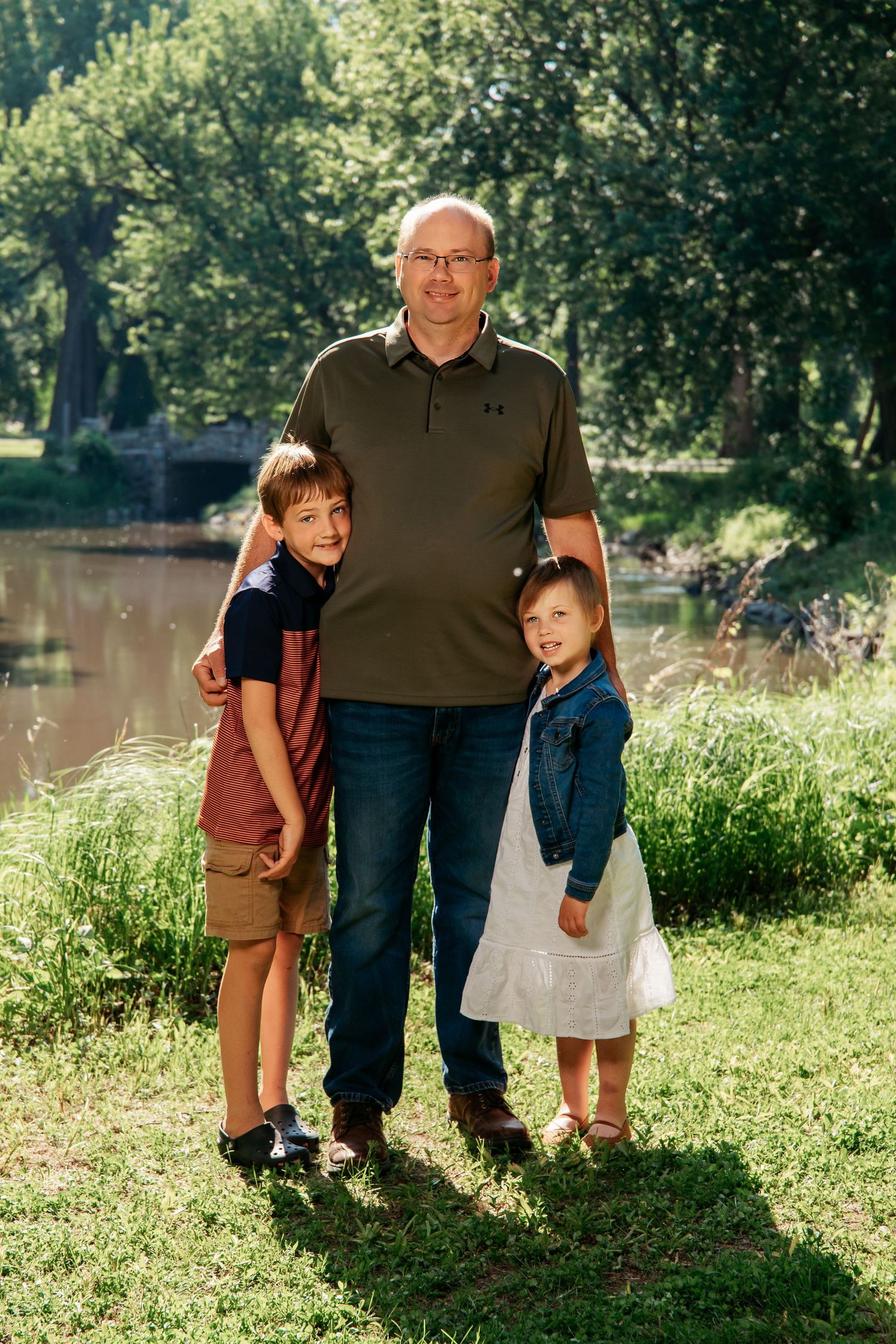 A family poses together on a sunny day by a tranquil pond in a lush green park setting.
