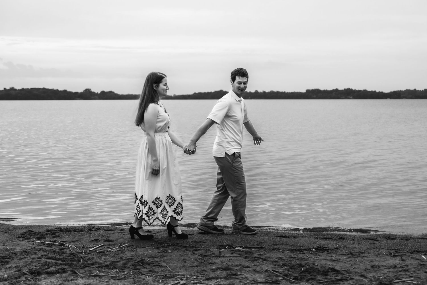 A couple in light colored outfits walks hand in hand along a lakeside in black and white photography.