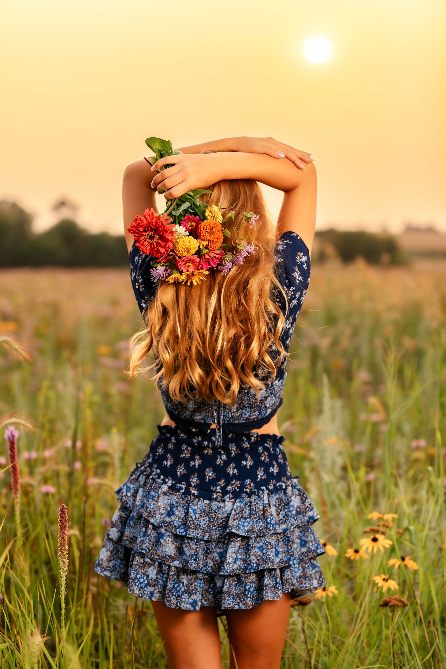 Back view of woman in floral dress holding colorful flowers in sunny meadow at sunset with golden sky.