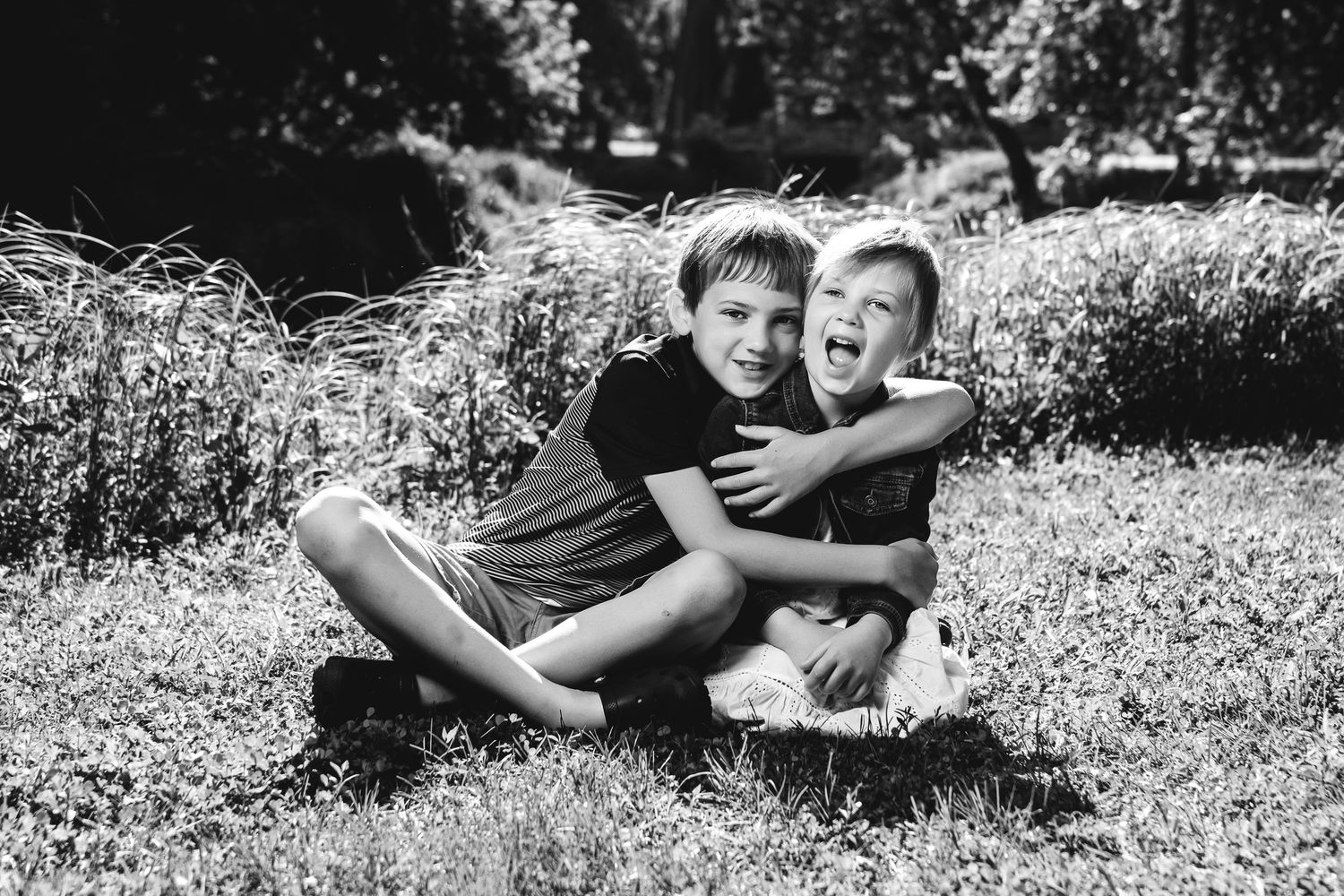 Two children laughing and playing together while sitting in grass on a sunny summer day in black and white.