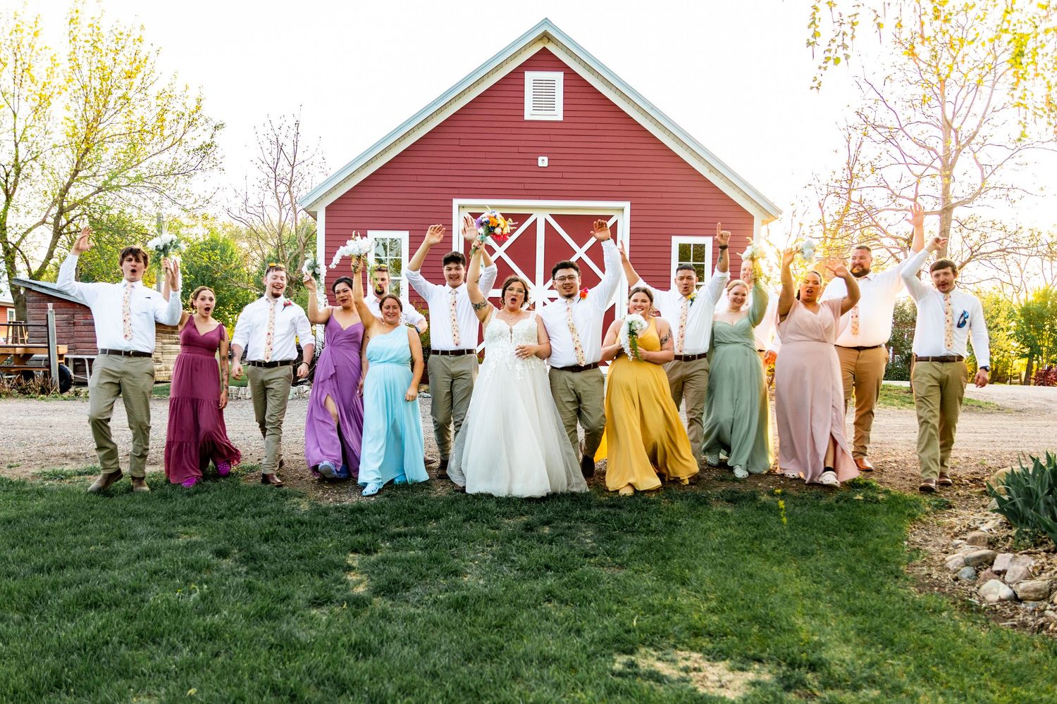 Wedding party jumps for joyful photo in front of red barn wearing pastel rainbow attire.