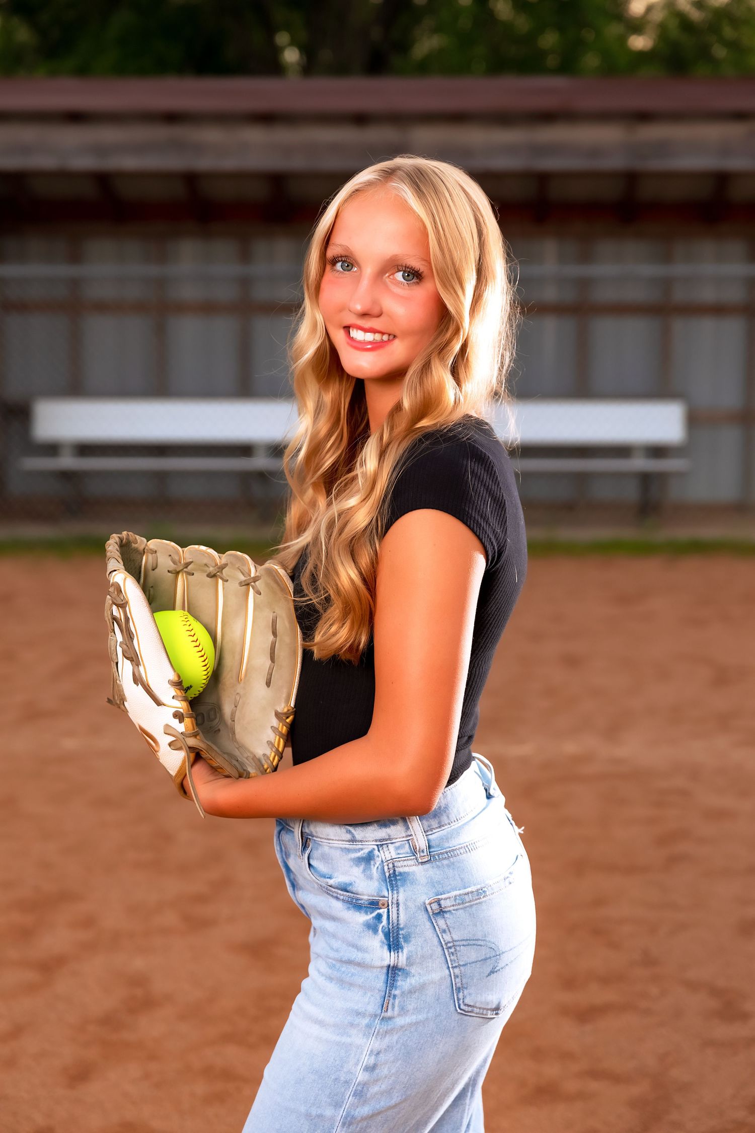 Senior photo shoot featuring a softball player in casual overalls on a dirt field at sunset.