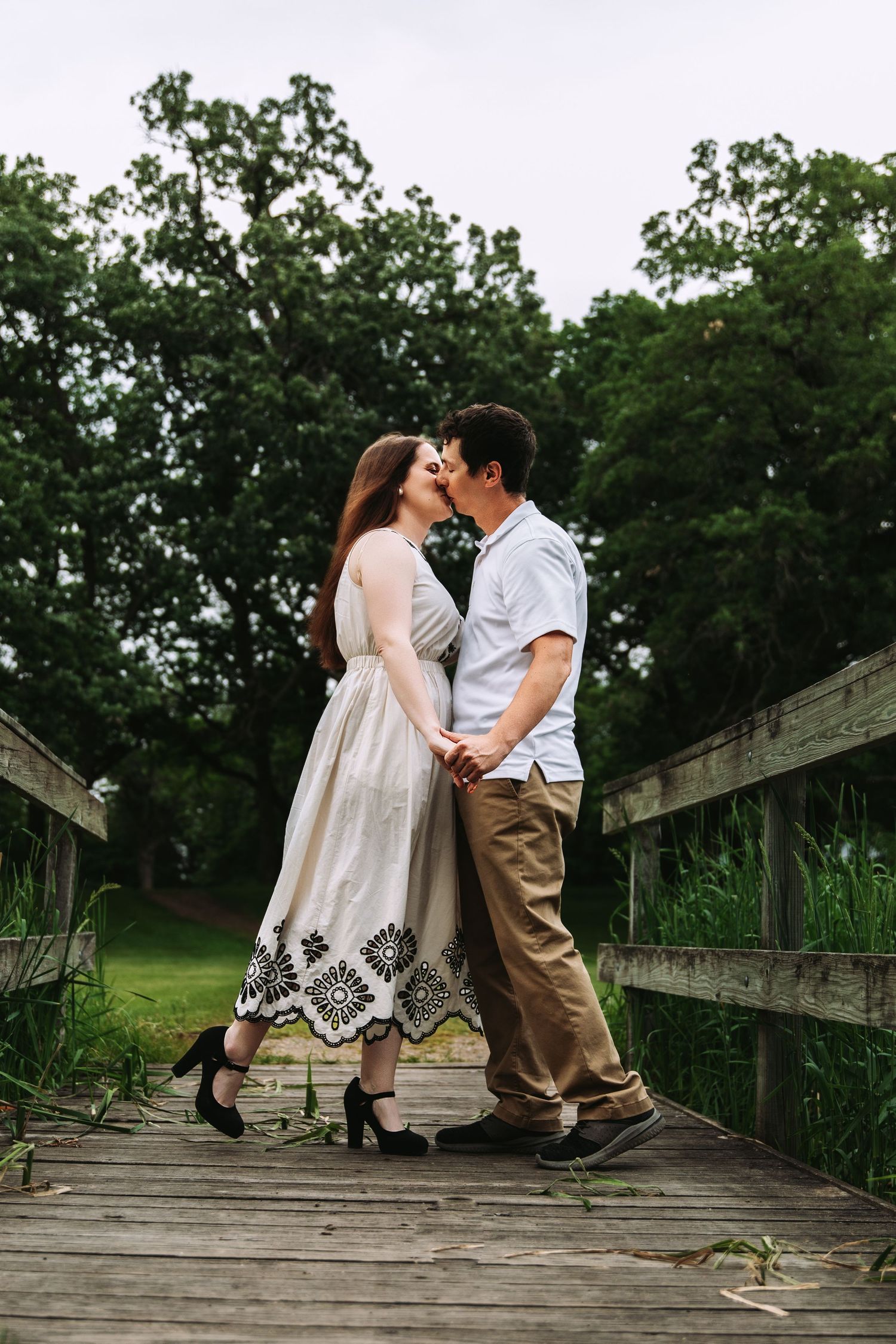 A romantic couple embraces and kisses on a wooden boardwalk surrounded by lush green trees in summer.