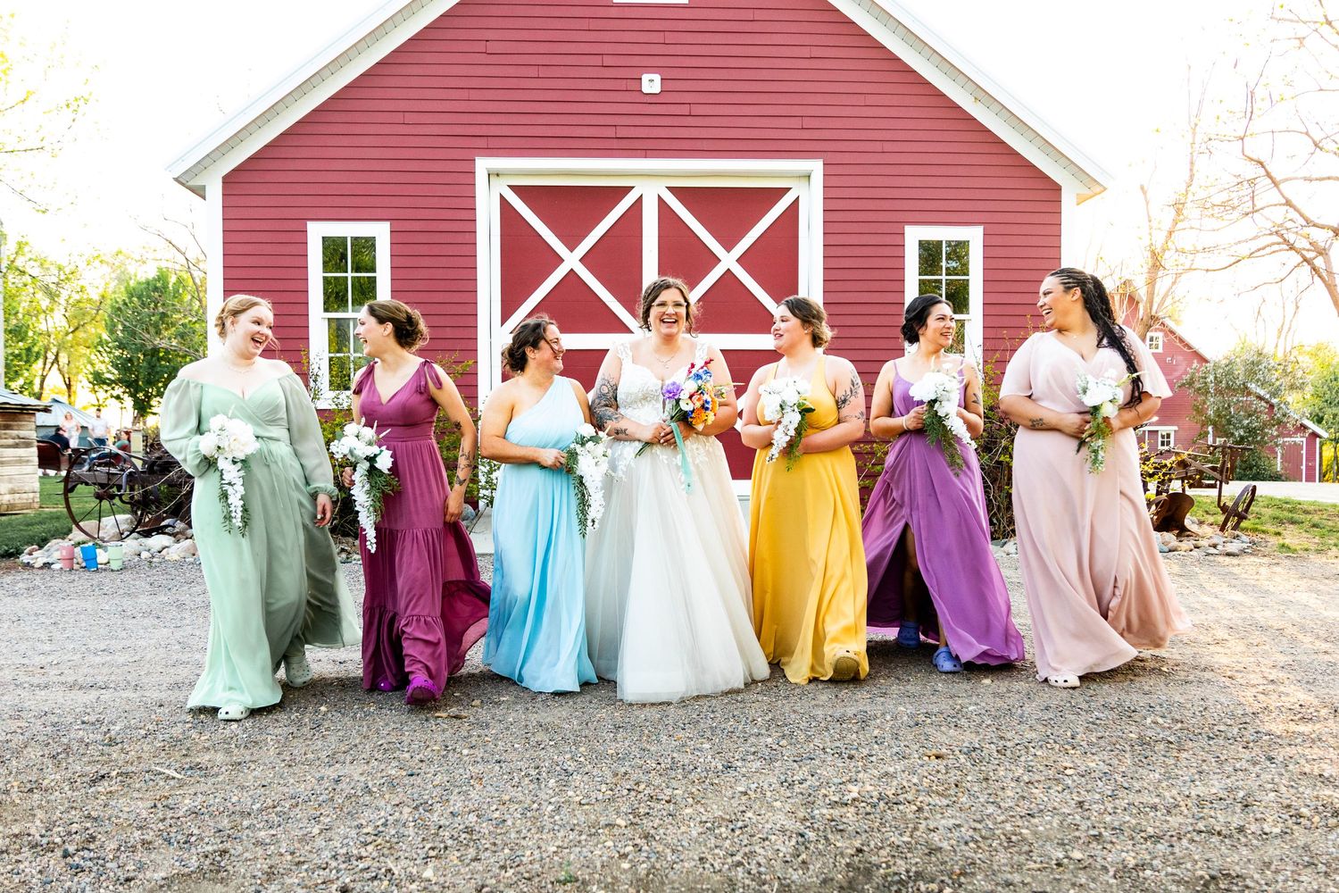 Wedding party poses in colorful rainbow bridesmaid dresses in front of a red barn on a sunny day.