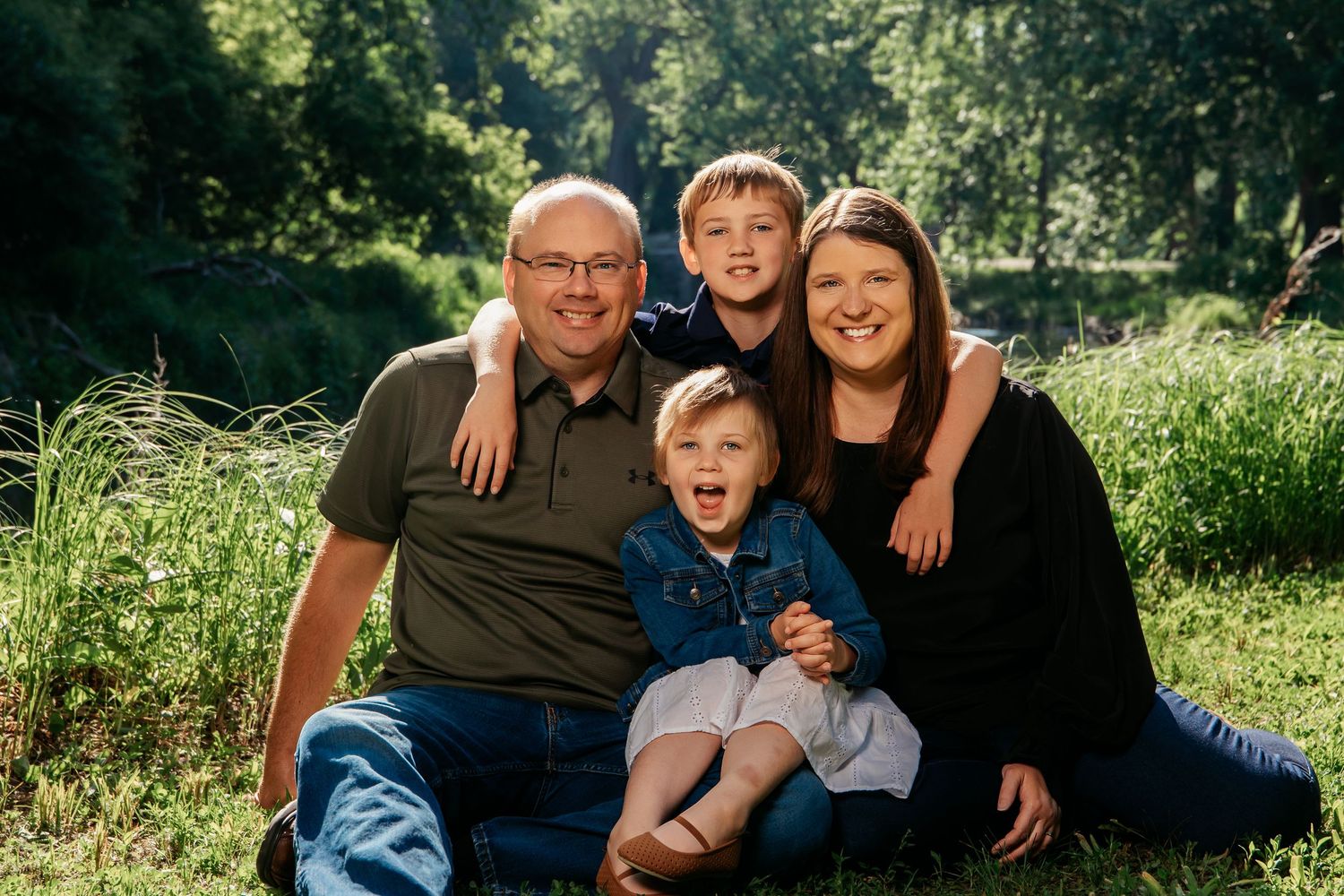 A happy family poses together outdoors on a sunny day in a grassy field surrounded by nature.