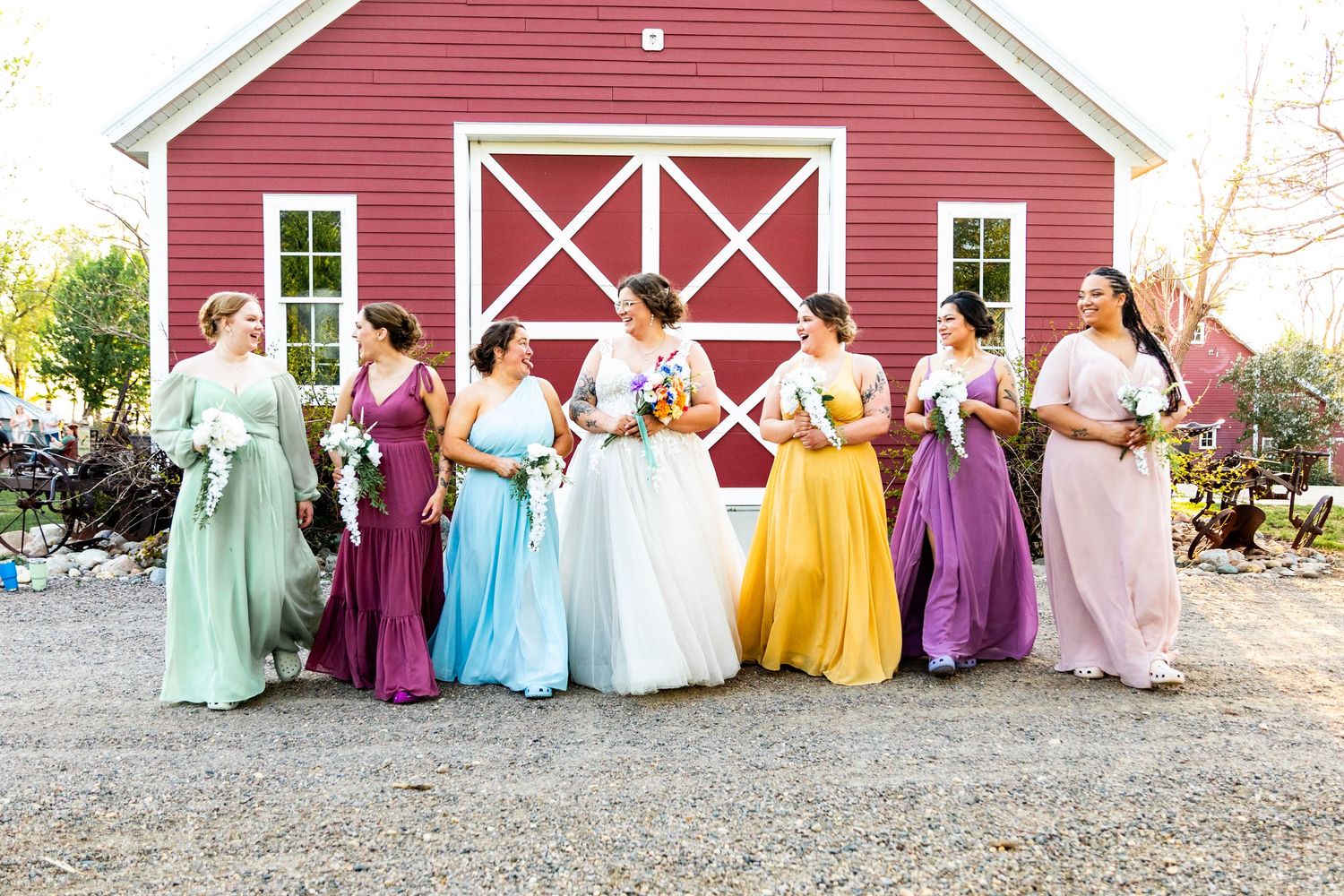 Wedding party in rainbow-colored dresses poses together in front of a bright red barn.