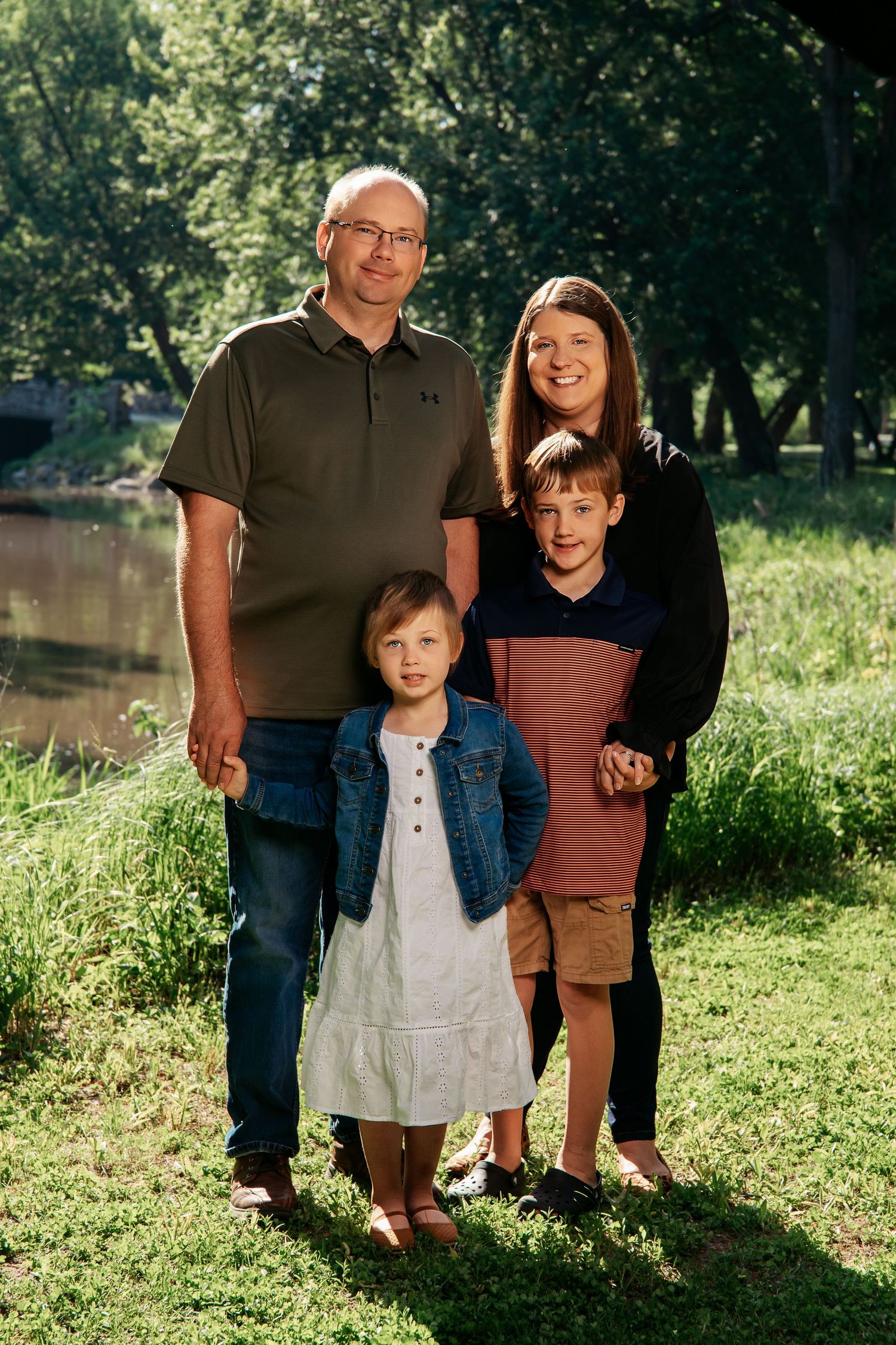 Family group poses together outdoors in a sunny green park setting with trees and water in the background.