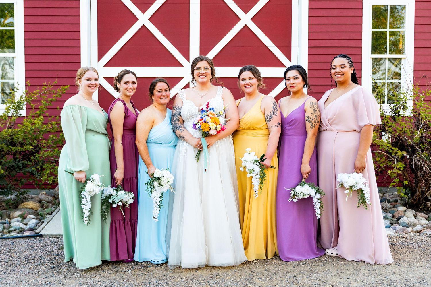 A rainbow of pastel bridesmaids dresses in front of a red barn wedding venue.