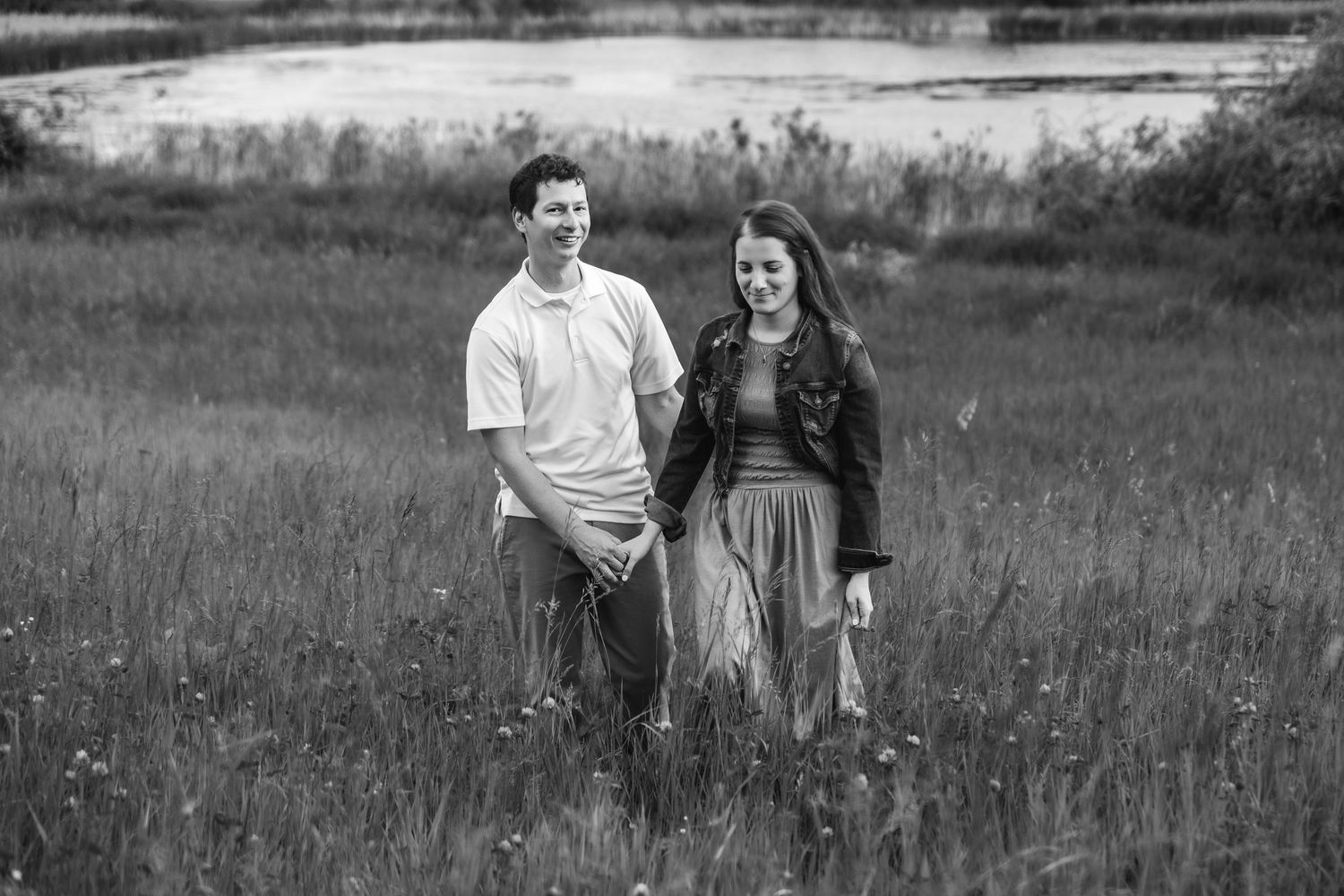 A couple stands together in a grassy meadow near a lake in this romantic black and white portrait.