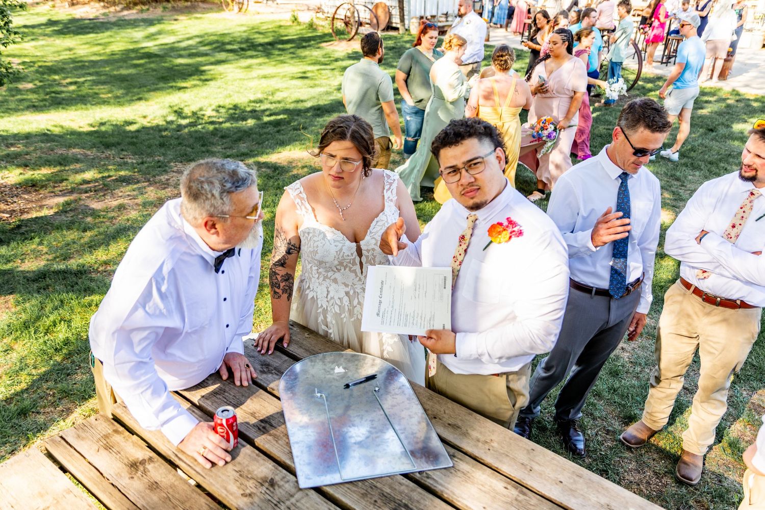 Wedding party and guests mingle at outdoor wooden picnic tables during a casual celebration.