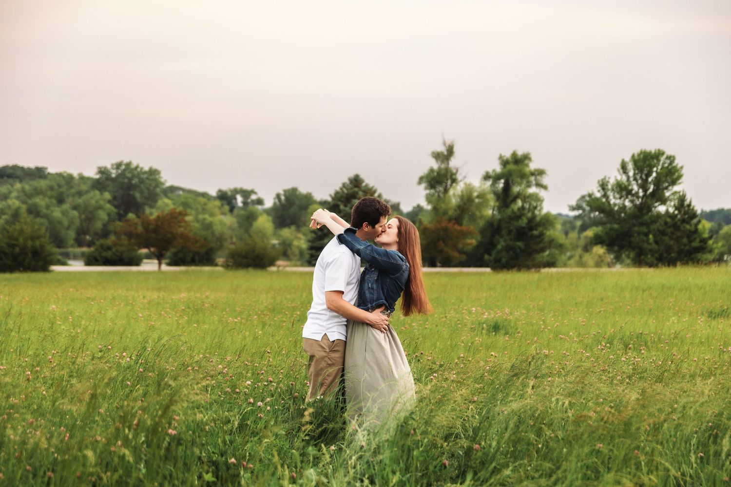 A couple embraces in a sunlit meadow surrounded by tall grasses and green countryside.