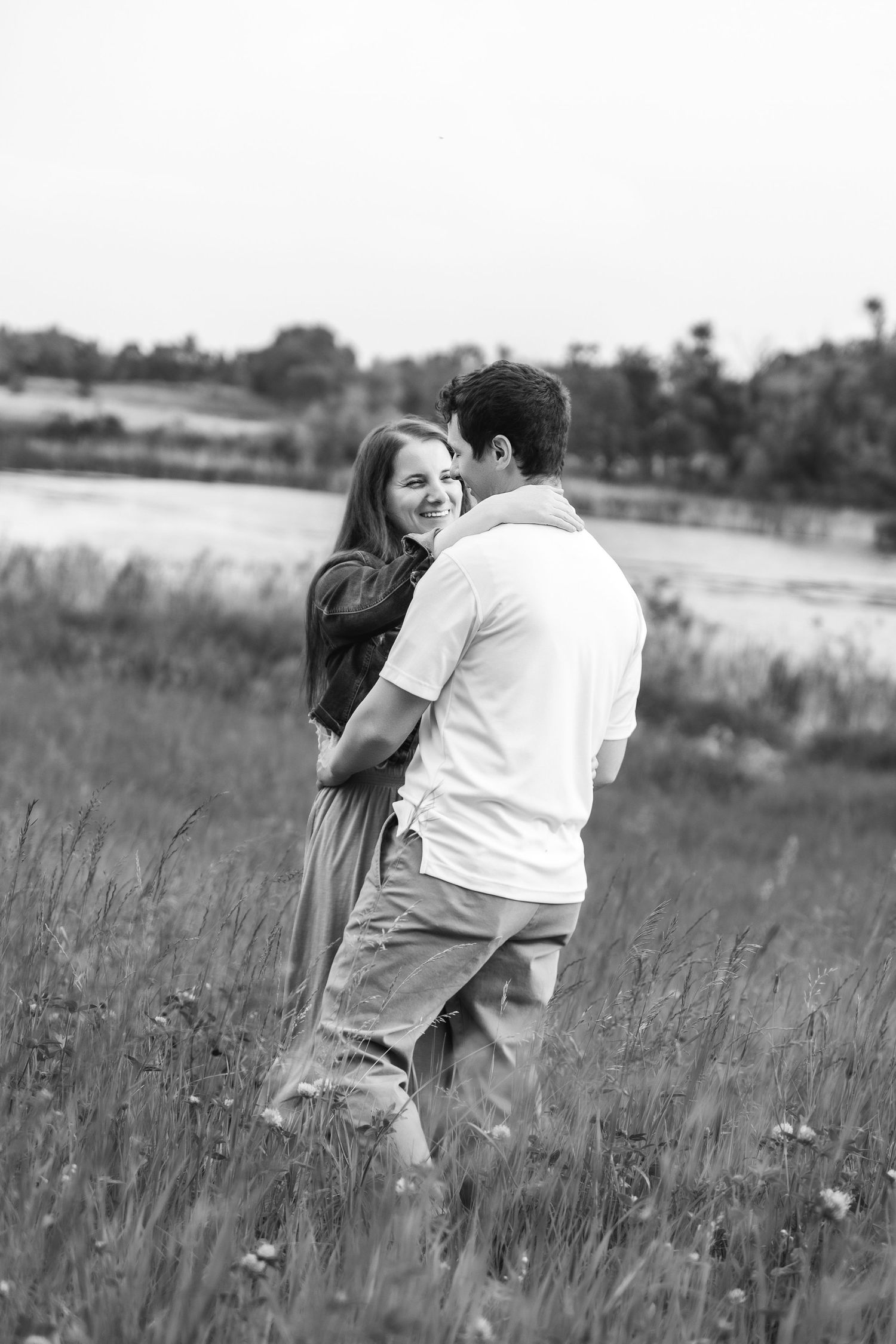 A romantic couple embraces in a serene meadow with wildflowers and tall grass in black and white.