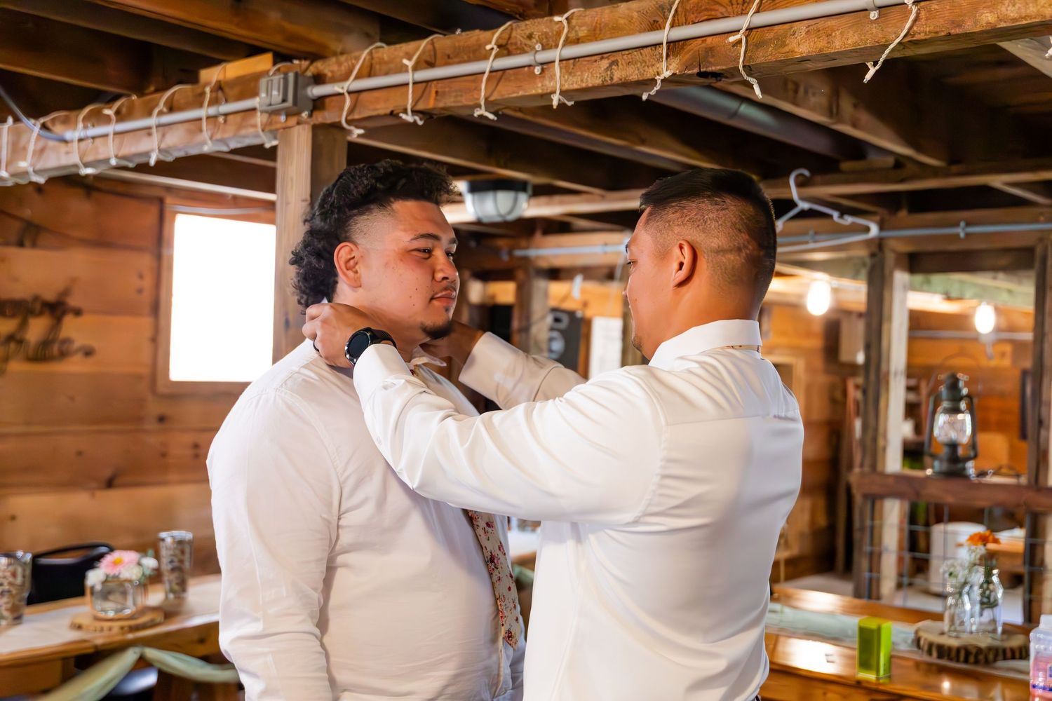 Two people in white shirts help each other with formal attire adjustments in rustic wooden room.