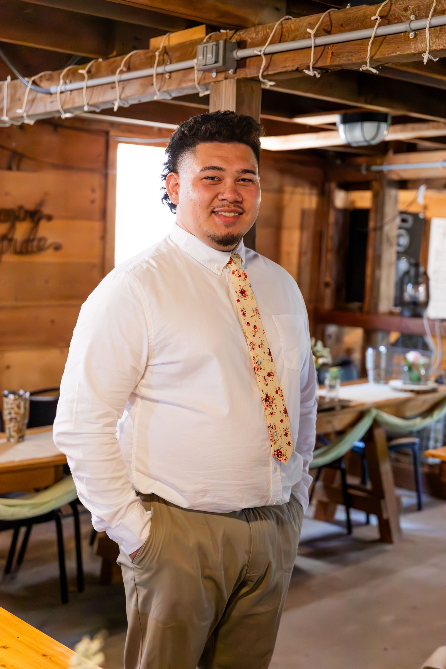 Professional dressed in a white dress shirt and patterned tie stands in a rustic restaurant interior with wooden walls.