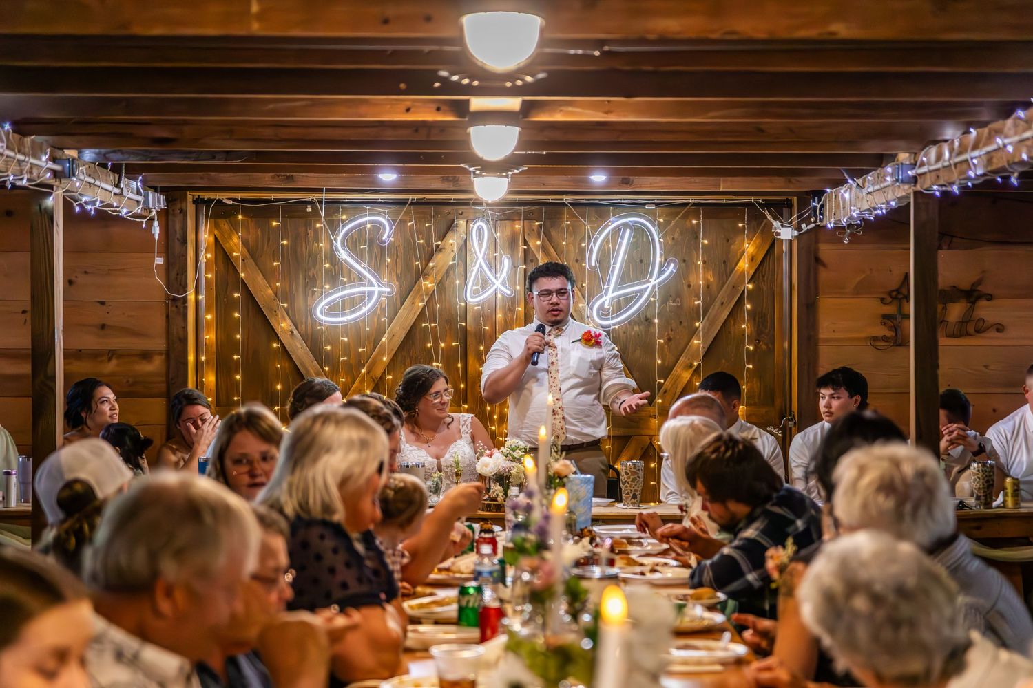 Wedding reception dinner in rustic wooden venue with glowing S&D initials and string lights on walls.