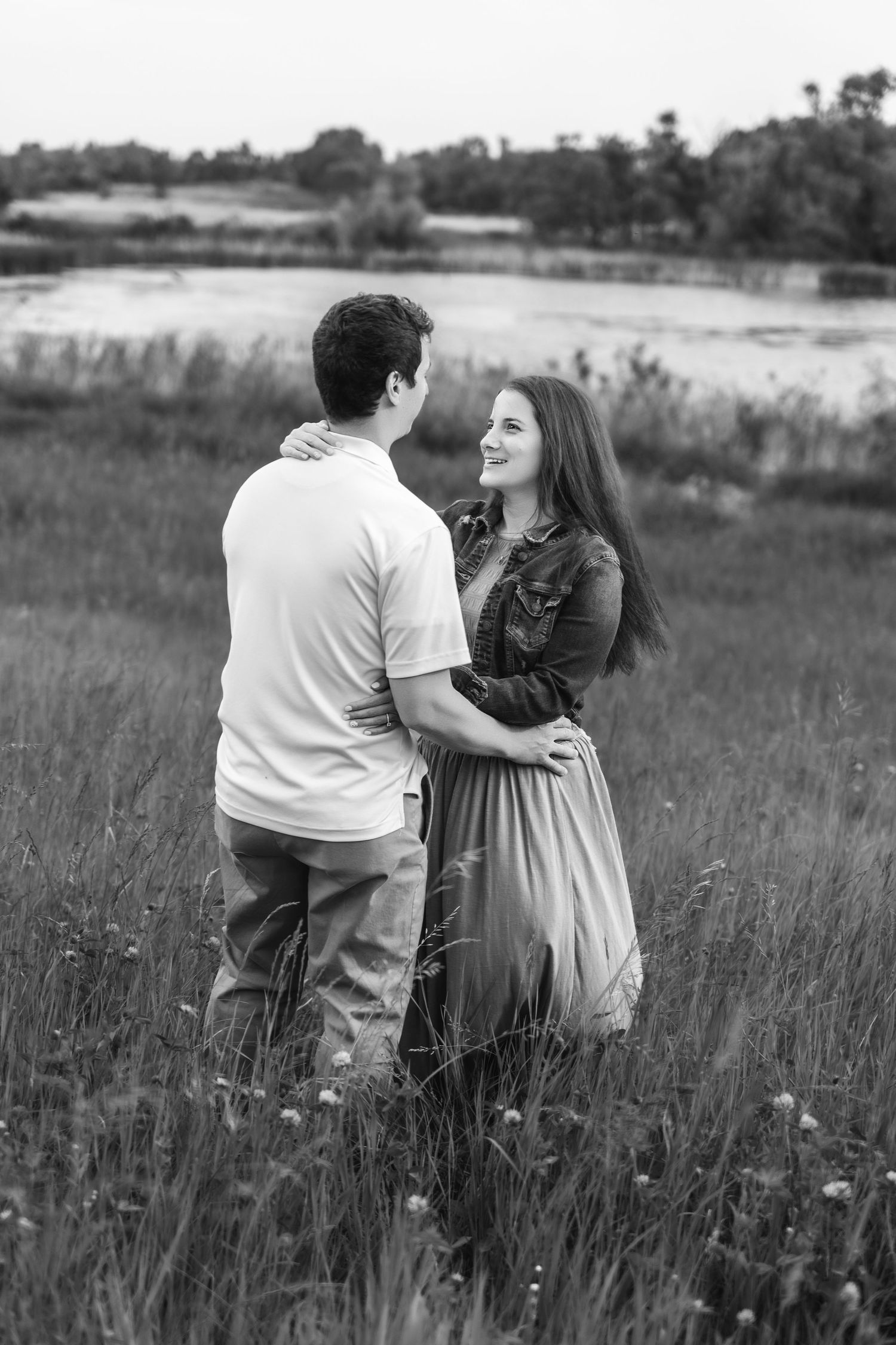 A romantic couple embraces while standing in a wildflower meadow near a lake in black and white photography.