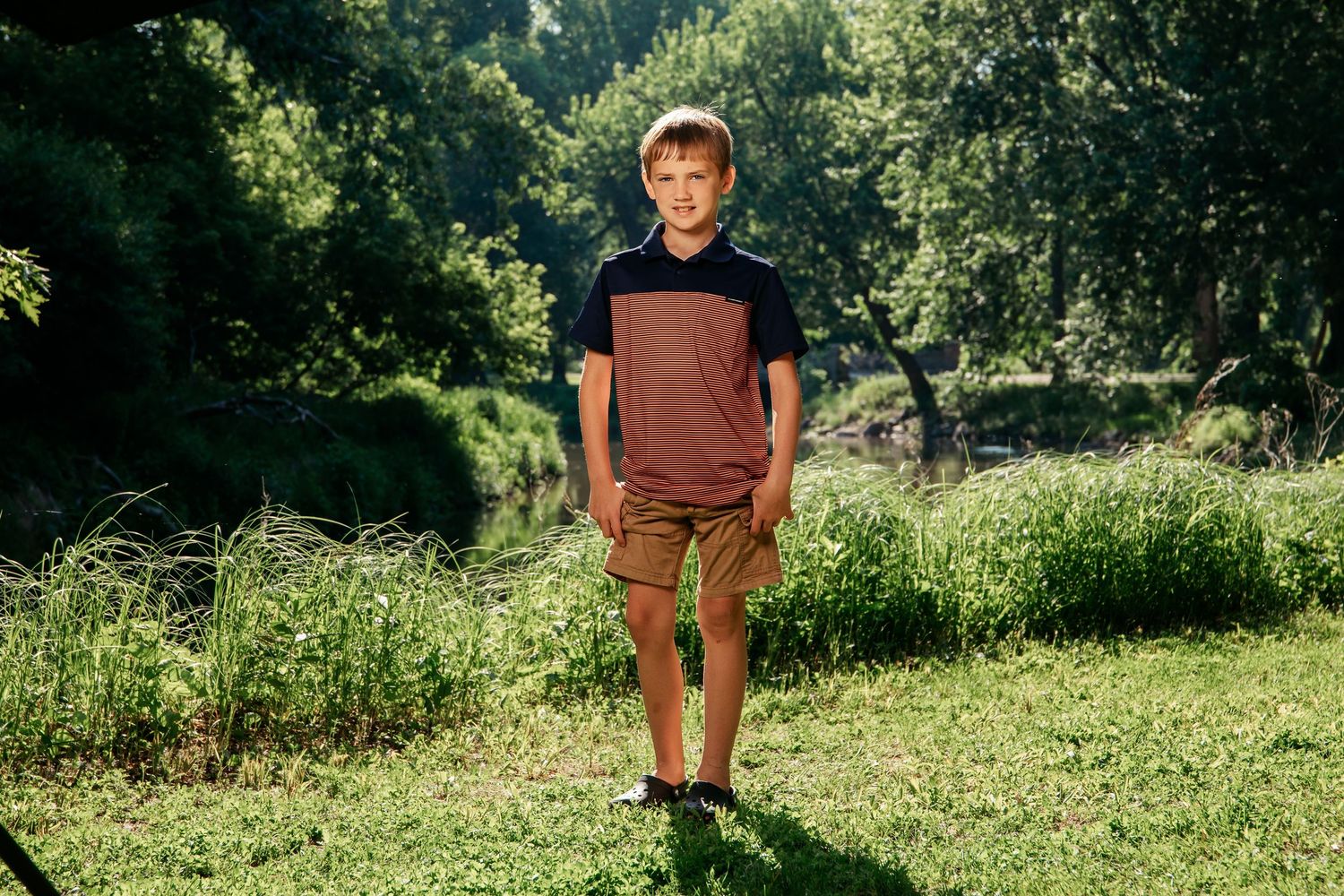 A person wearing brown shorts and navy t-shirt stands in sunlit grassy field with trees in background.