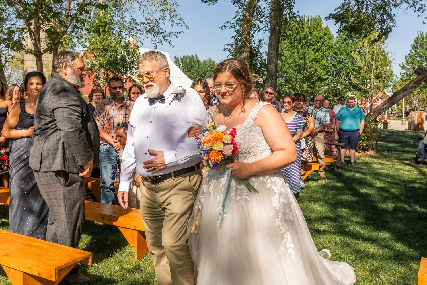 Outdoor garden wedding ceremony with guests seated on wooden benches under trees.