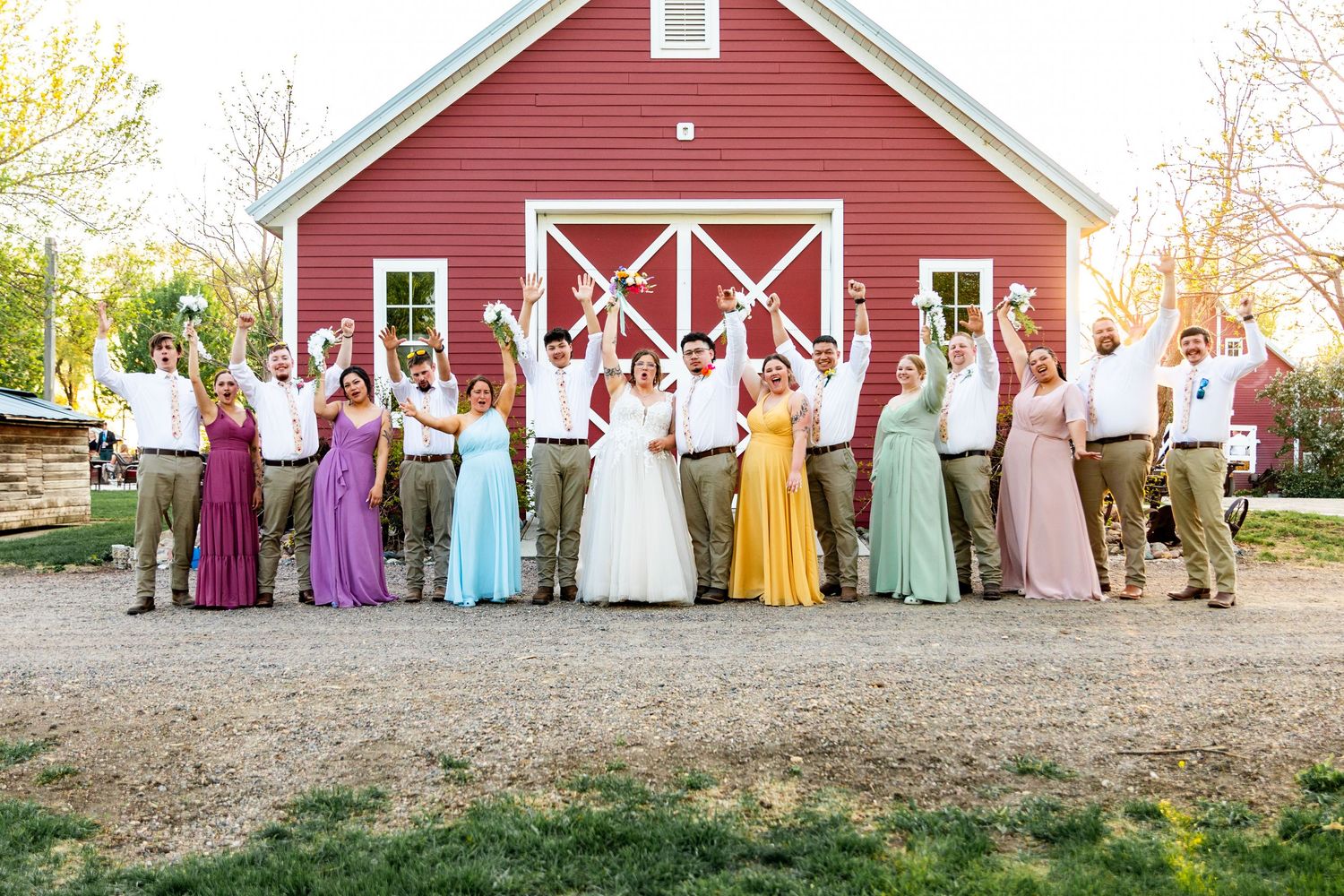Large wedding party in pastel dresses and formal wear gathered by a red barn.