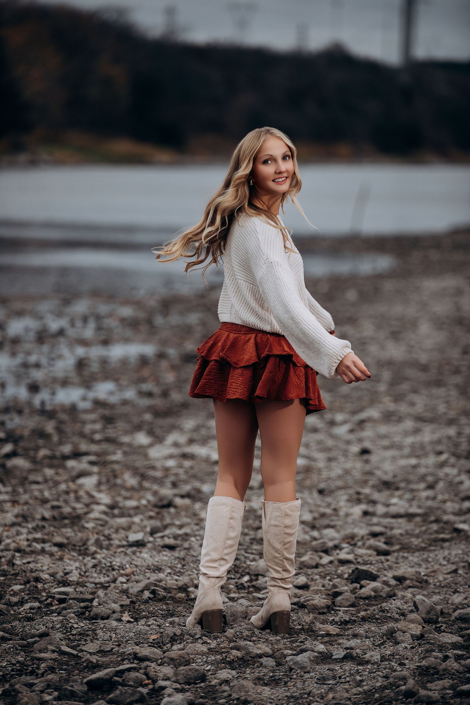 Woman in sweater and ruffled skirt twirls on rocky beach wearing tall white boots.