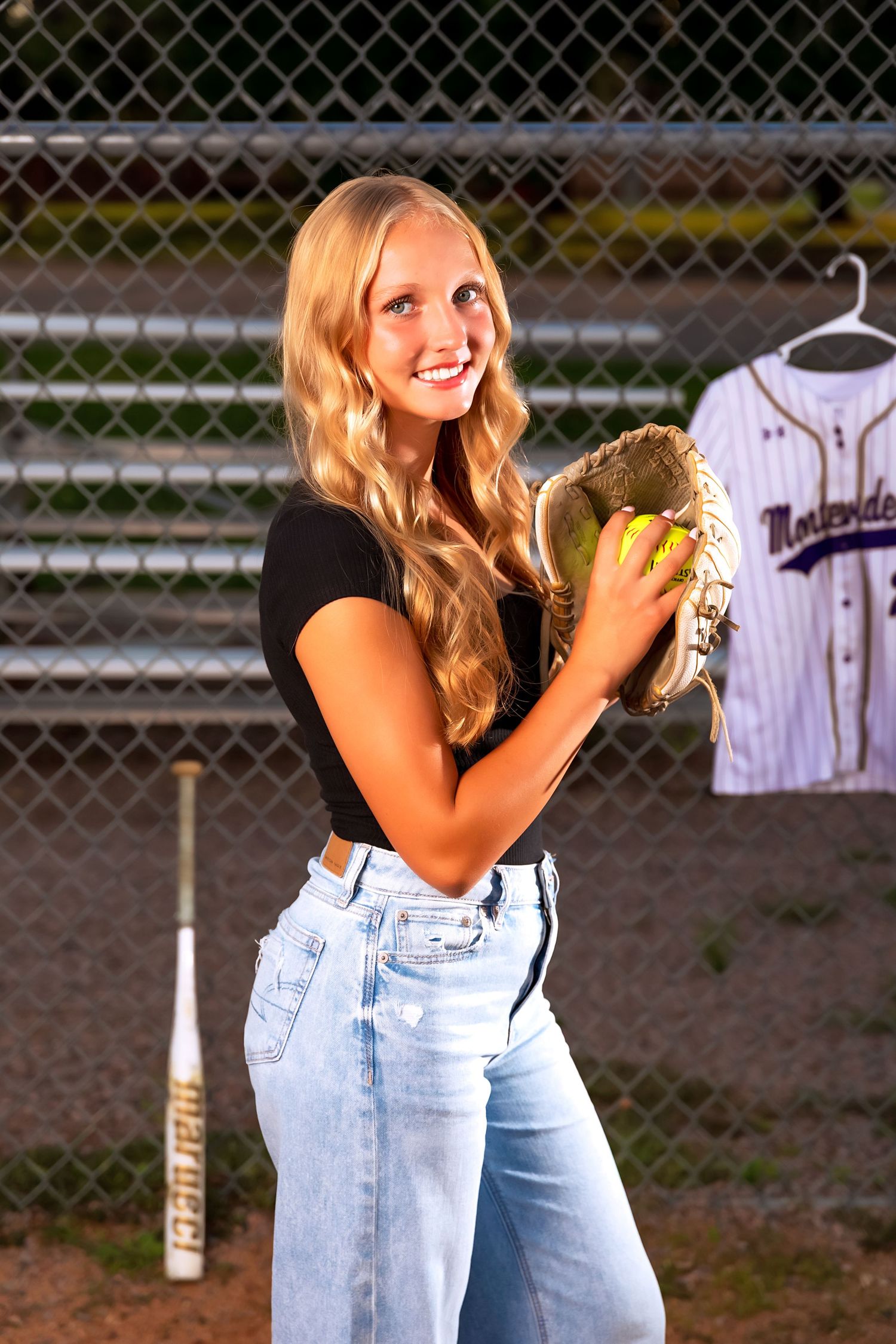Student athlete poses with baseball glove in front of dugout and baseball jersey.