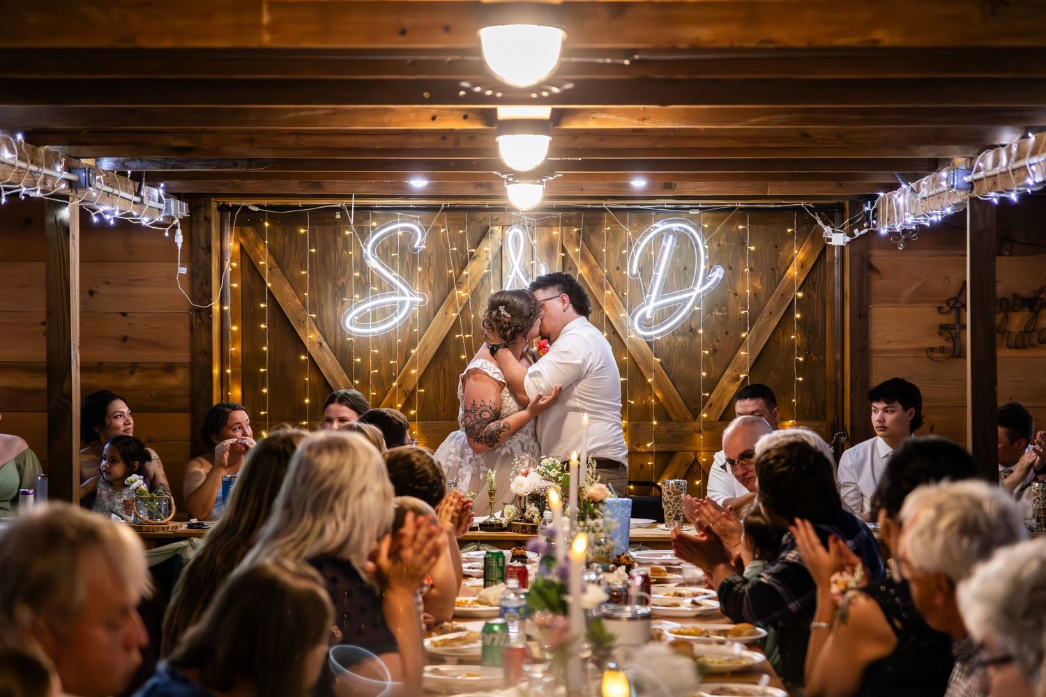 A romantic wedding reception scene in a rustic wooden barn with twinkle lights and guests seated at long tables.