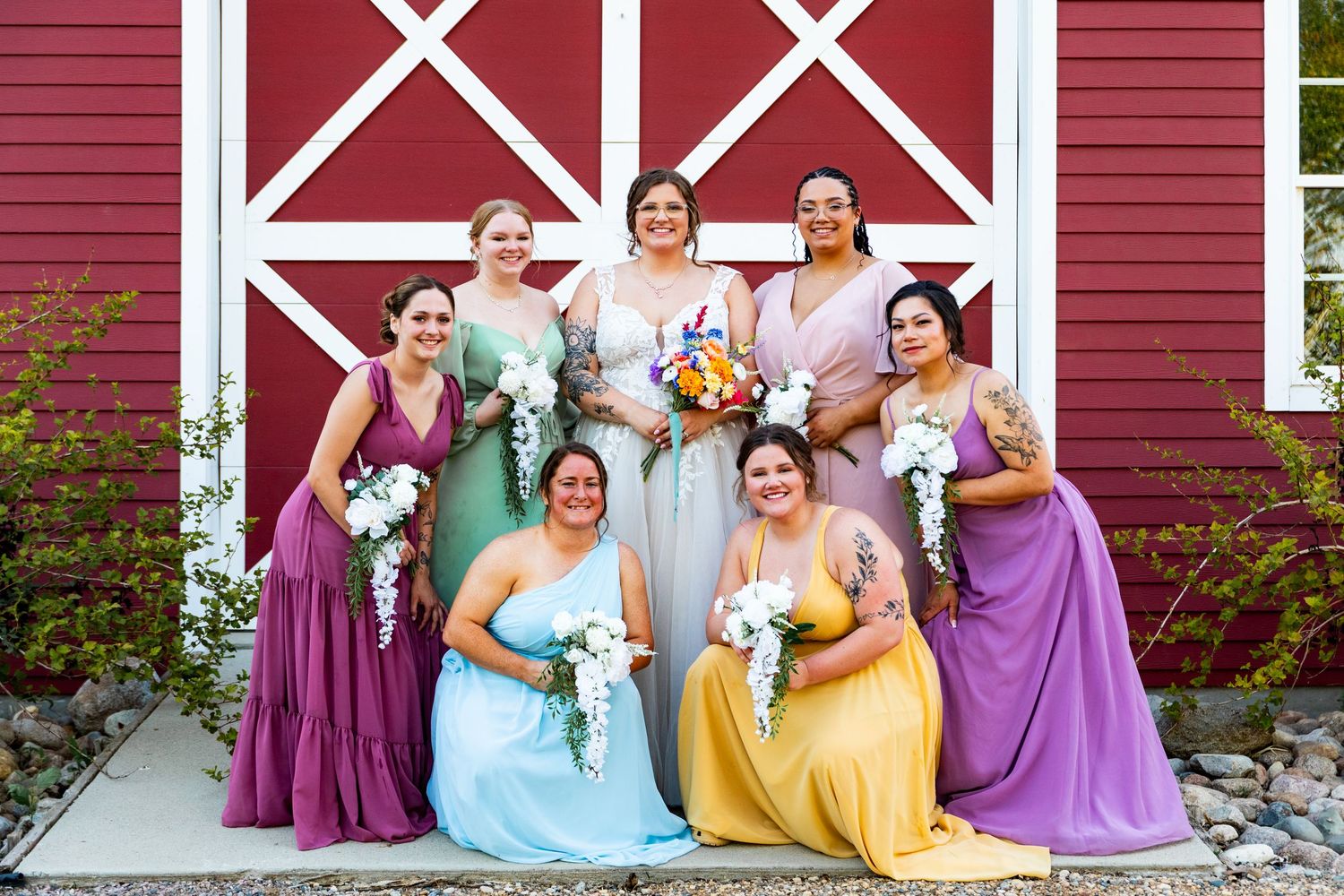Colorful wedding party poses in front of a red barn wearing dresses in shades of purple, yellow and blue.