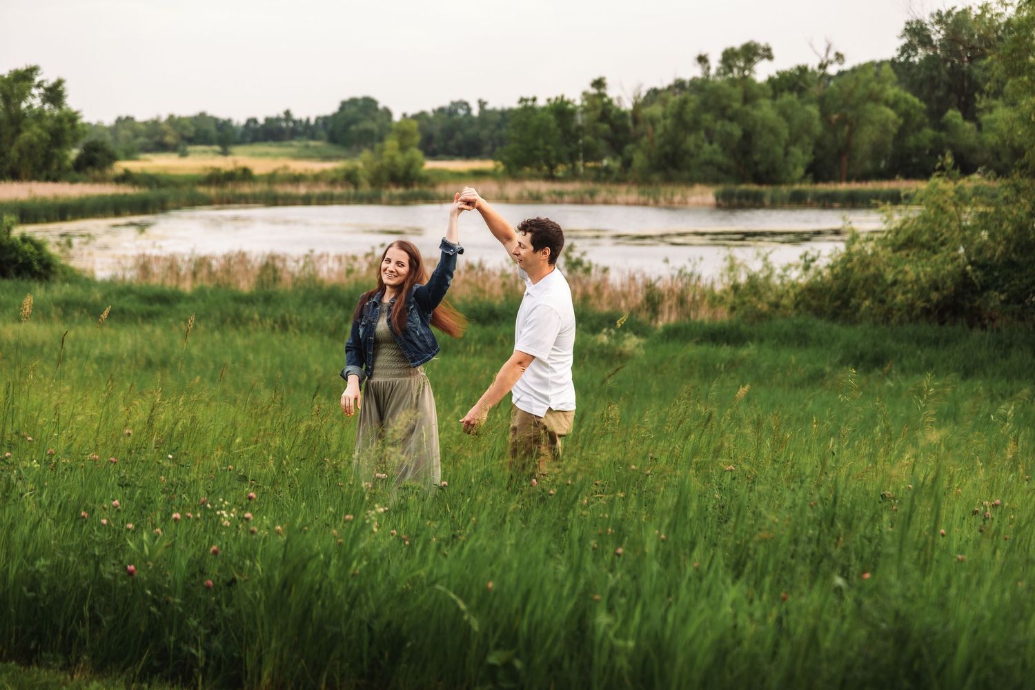 A couple dancing and twirling in a lush green meadow near a serene pond on a summer day.
