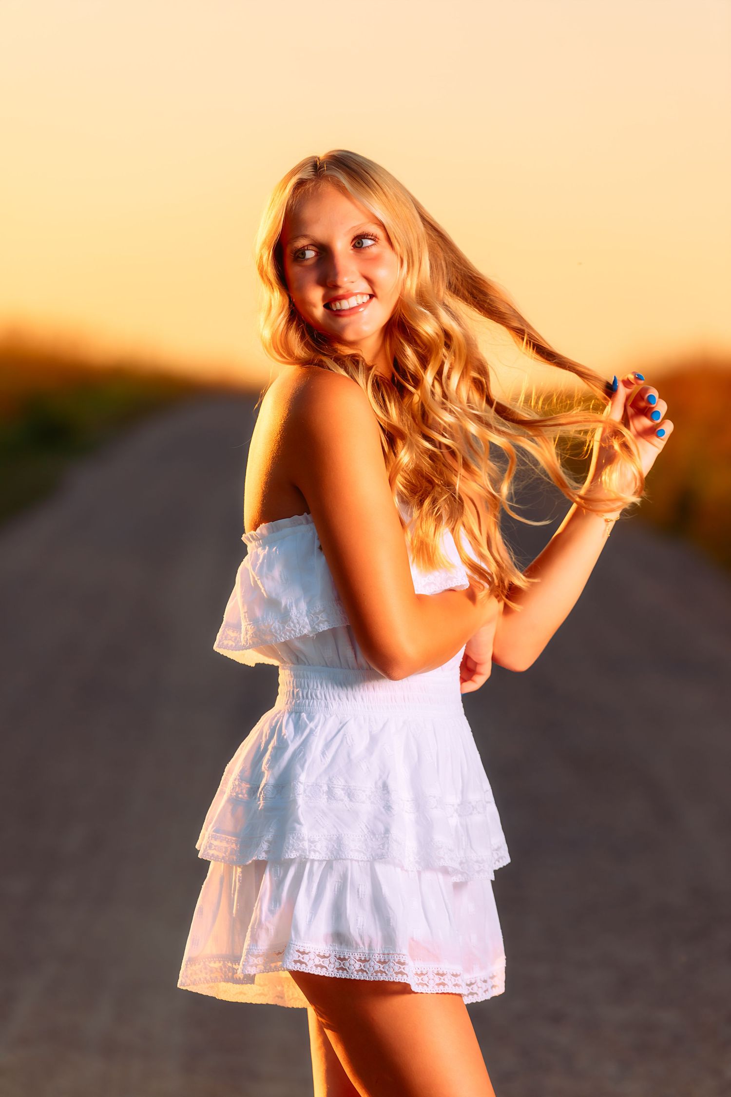 Young woman in white summer dress poses playfully on empty road during golden sunset.