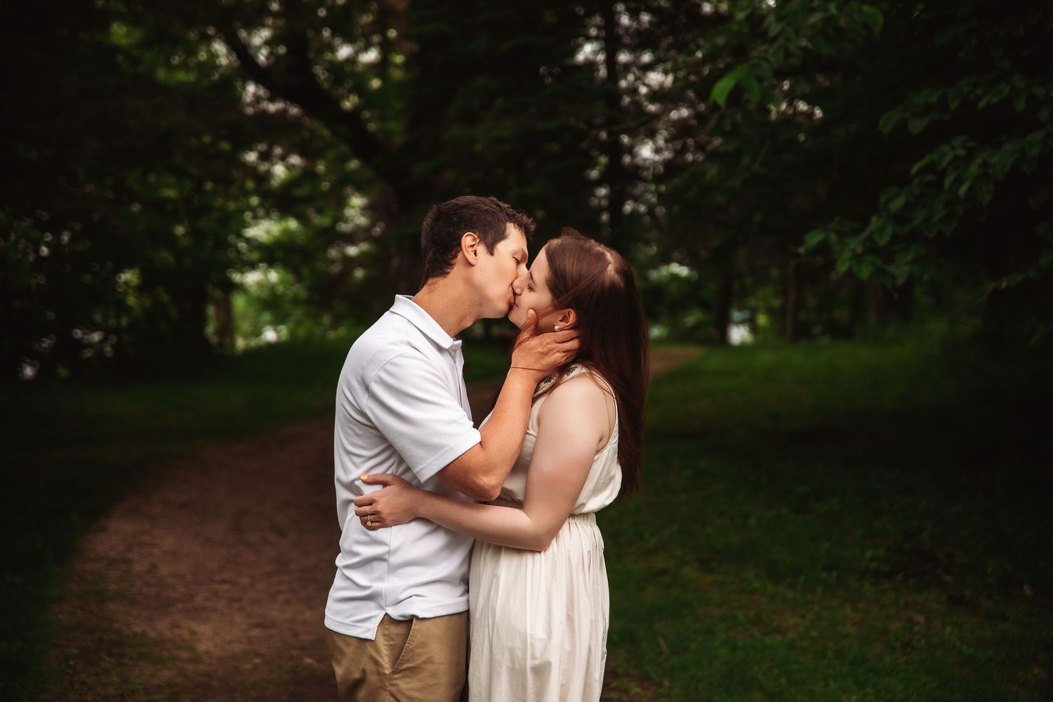 A couple in casual attire shares a romantic kiss on a tree-lined path.