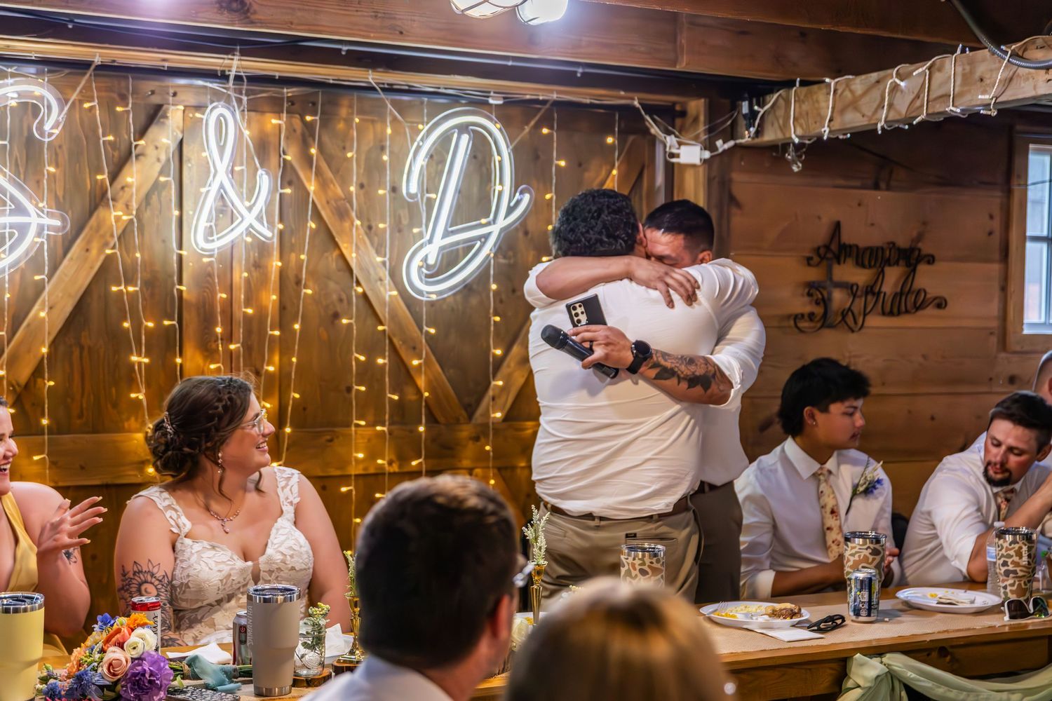 Wedding reception celebration inside rustic barn with decorative string lights and neon signage.