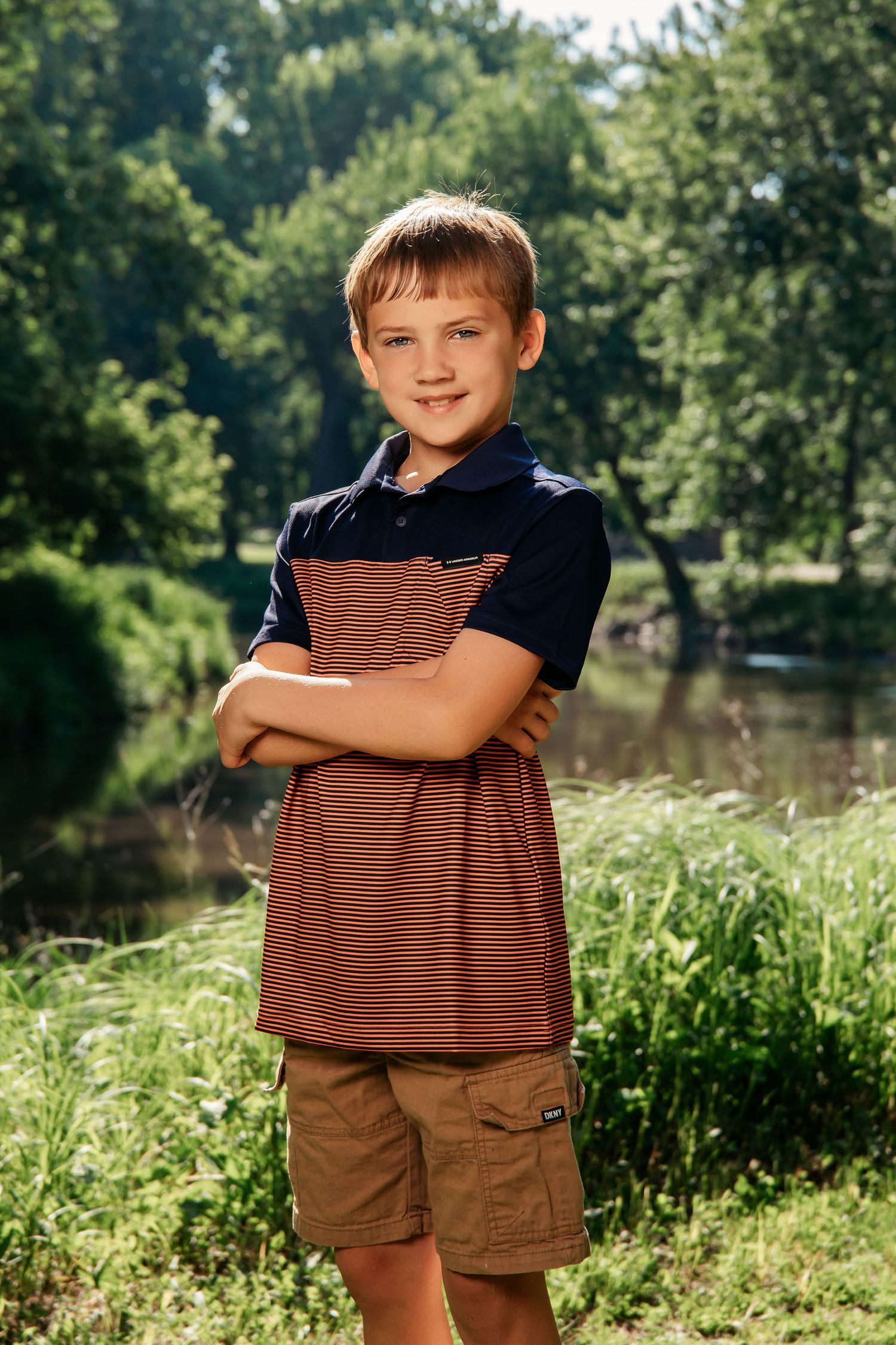 Portrait of a youth standing by a scenic pond wearing a striped navy and brown polo shirt and khaki cargo shorts.