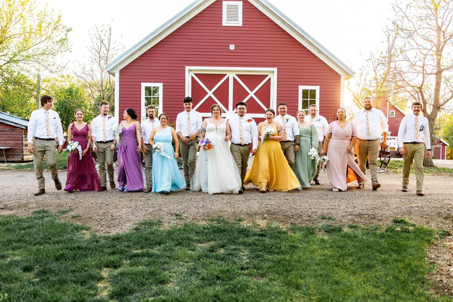 Wedding group photo featuring bridesmaids in rainbow-colored dresses against a red barn backdrop.