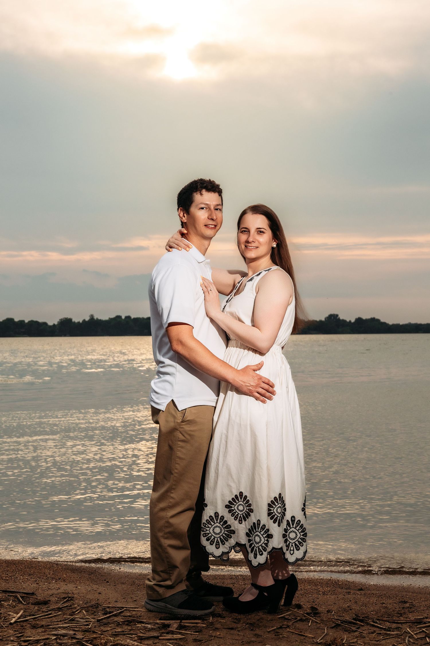 Romantic couple embracing on a beach at sunset with water and cloudy sky in the background.