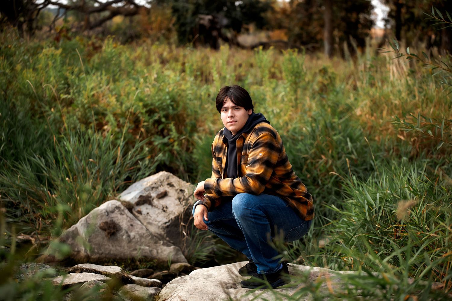 A person in striped sweater sits on rocks in a natural outdoor setting surrounded by tall grass and autumn foliage in Pipestone, Minnesota.