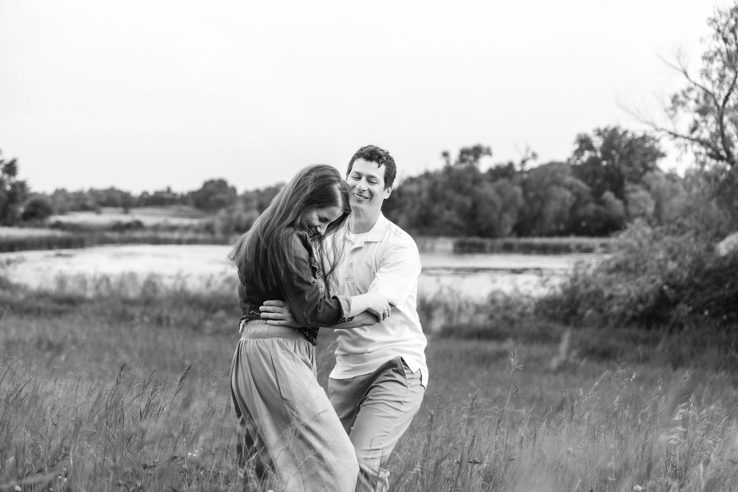 A couple embraces and laughs together while standing in a grassy meadow in black and white.