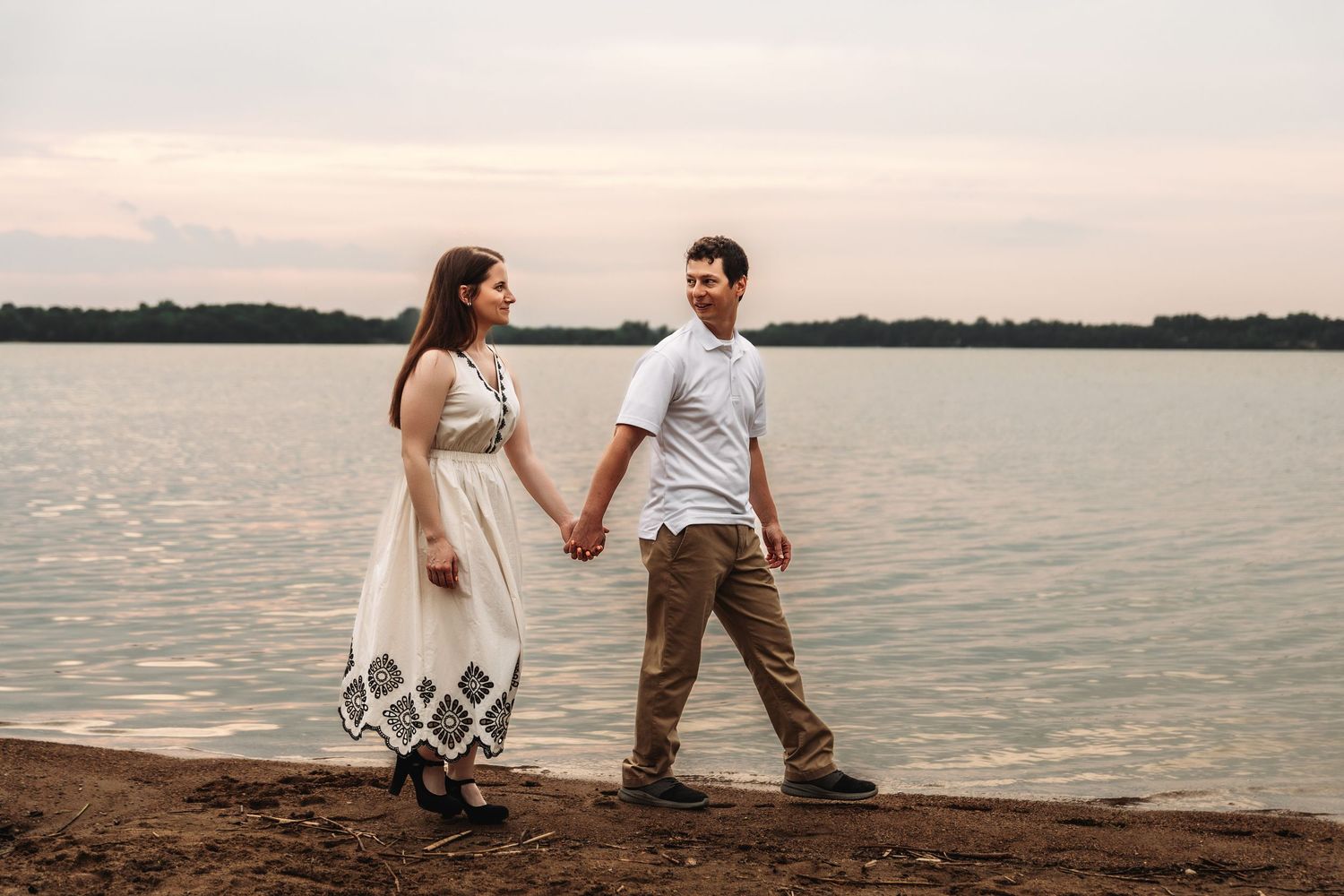 A couple stands holding hands at the edge of a serene lake during sunset.