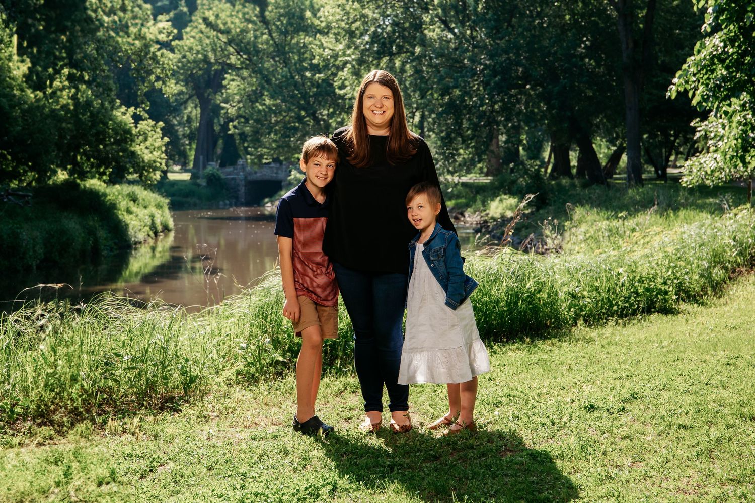 Three people stand together on a grassy riverbank in a sunlit park setting on a summer day.
