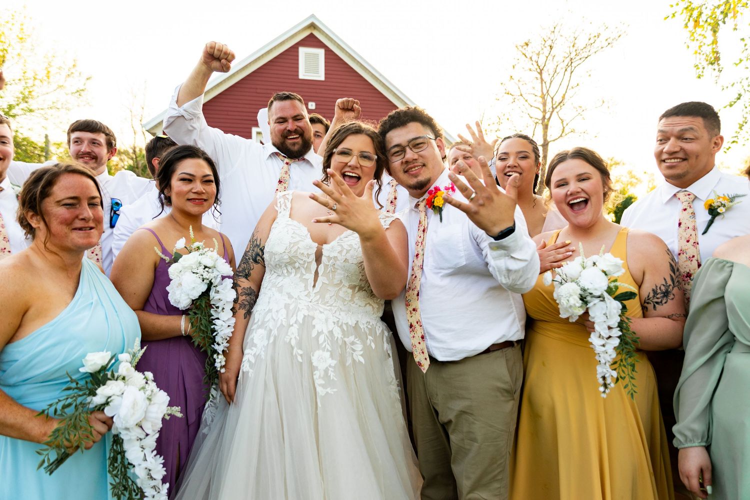 Joyful wedding party cheers together in front of red barn during outdoor celebration.