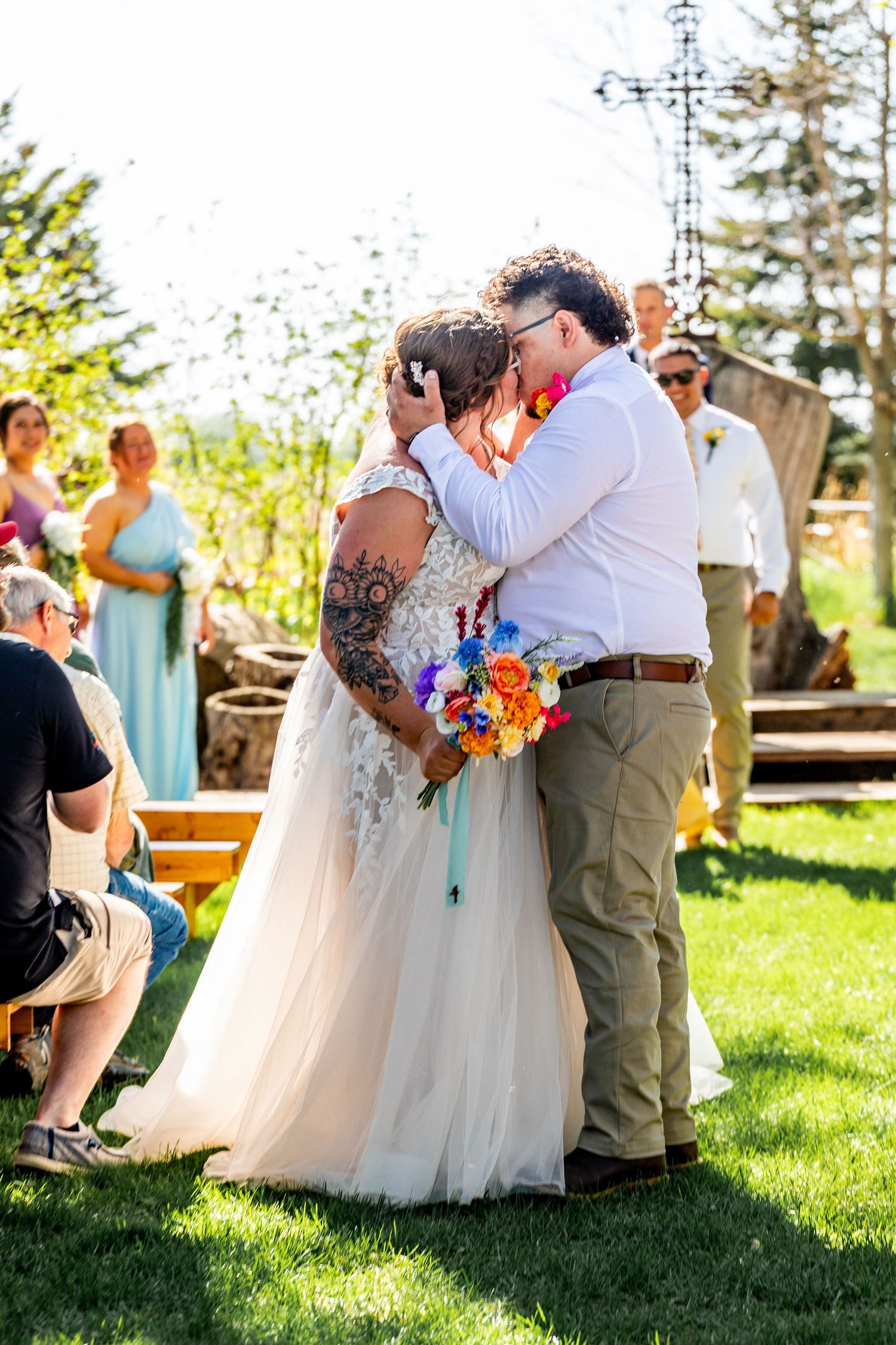 A couple shares their first kiss during an outdoor wedding ceremony with colorful flowers and guests looking on.