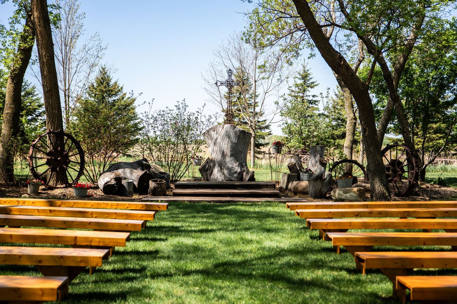 Wooden benches arranged in rows on a grassy lawn create an outdoor ceremony space under tall trees.
