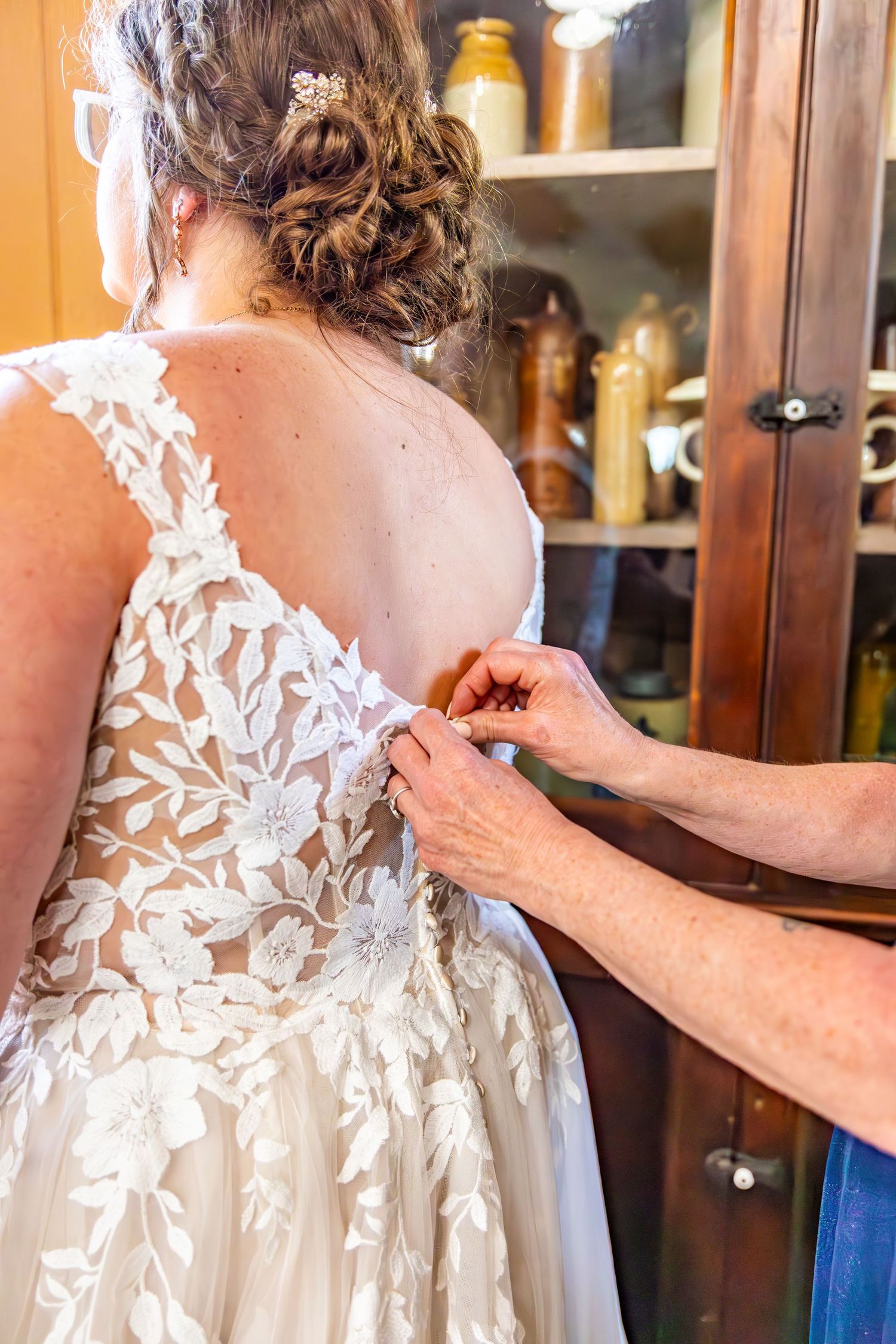 Close-up detail of a wedding dress showing intricate white floral lace patterns and delicate buttons being fastened.