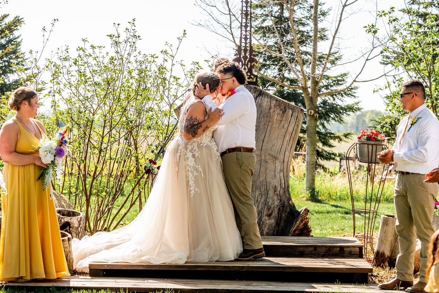 A romantic wedding ceremony takes place outdoors on a rustic wooden platform surrounded by spring greenery.