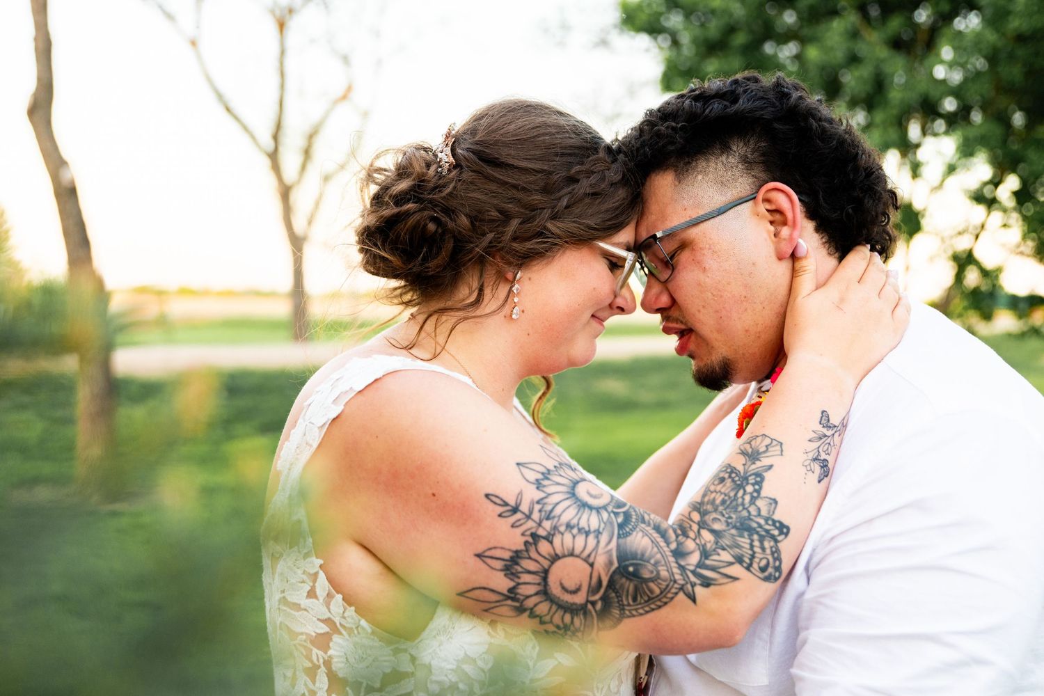 A romantic outdoor photo showing two people in white clothing with visible tattoos embracing in a sunlit natural setting.